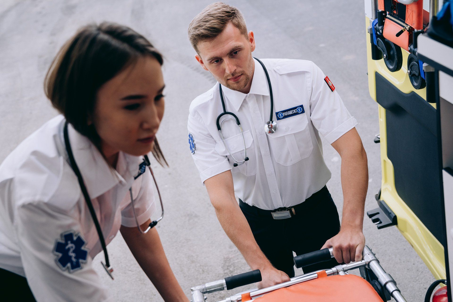 Two paramedics loading a stretcher into an ambulance; one male, one female, both wearing white uniforms, focused.
