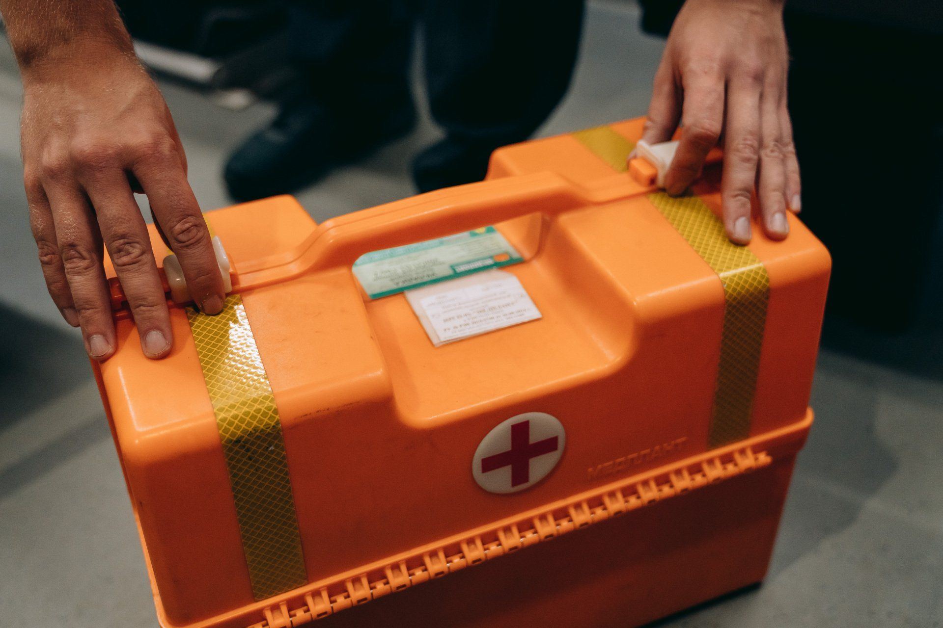 Mani che tengono un kit di pronto soccorso arancione con una croce bianca, con la maniglia rivolta verso l'alto, pronto per essere aperto.