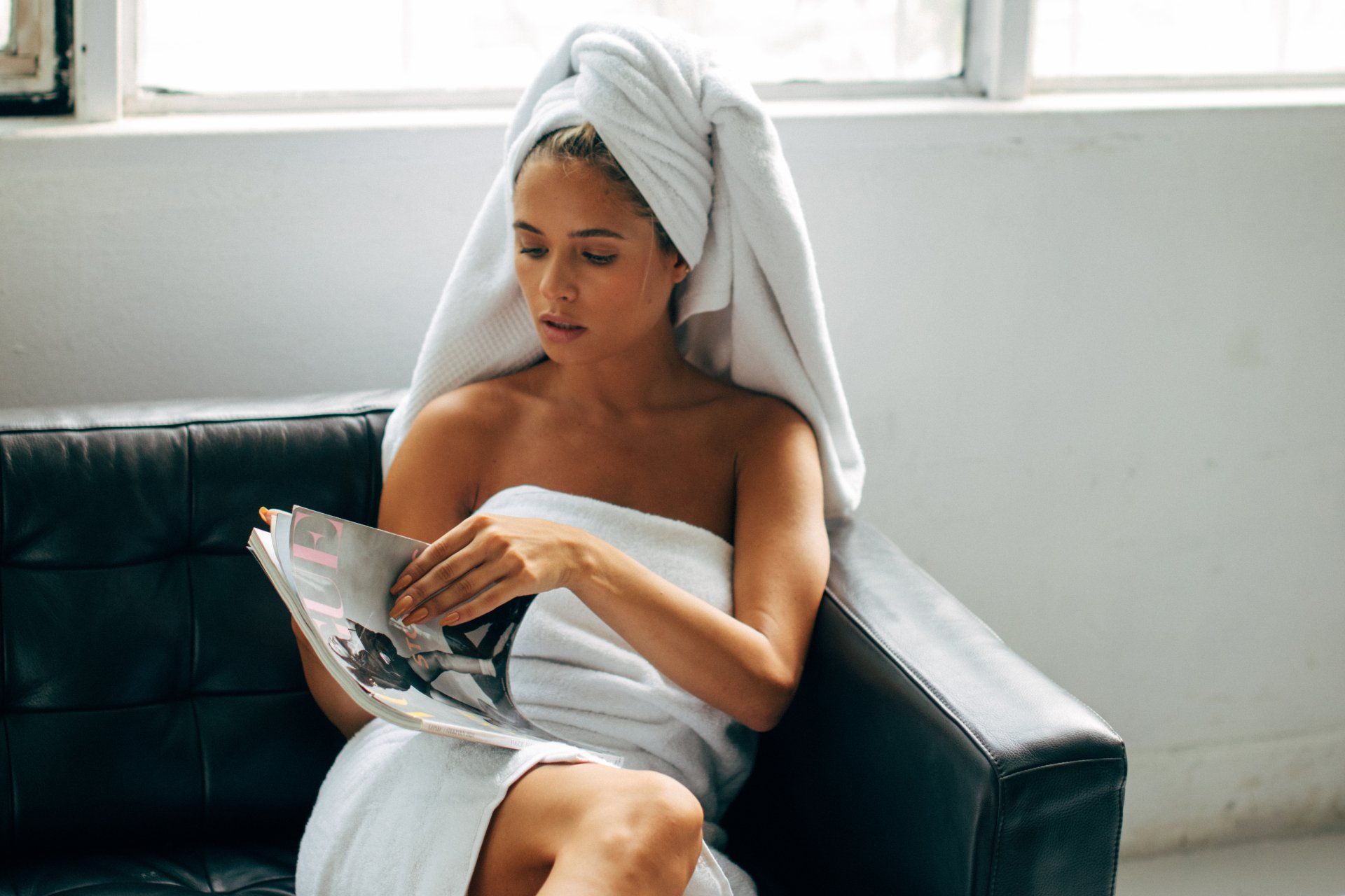 A woman wrapped in a towel is sitting on a couch reading a magazine.