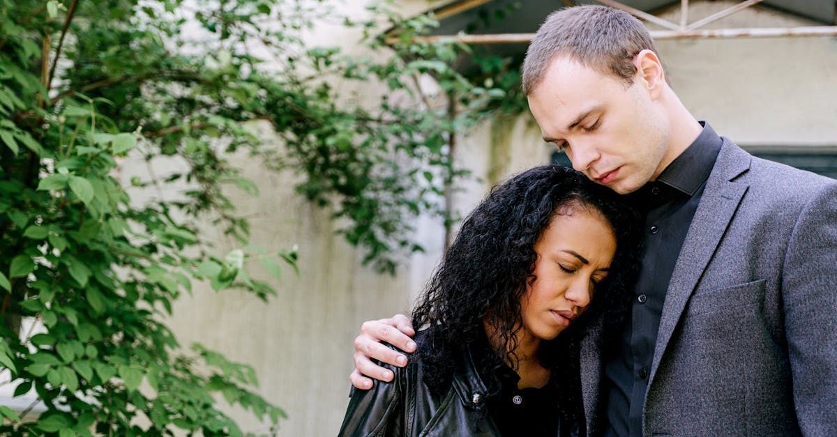 A person in a blazer comforts another person with curly hair who is leaning against their shoulder in an outdoor setting.
