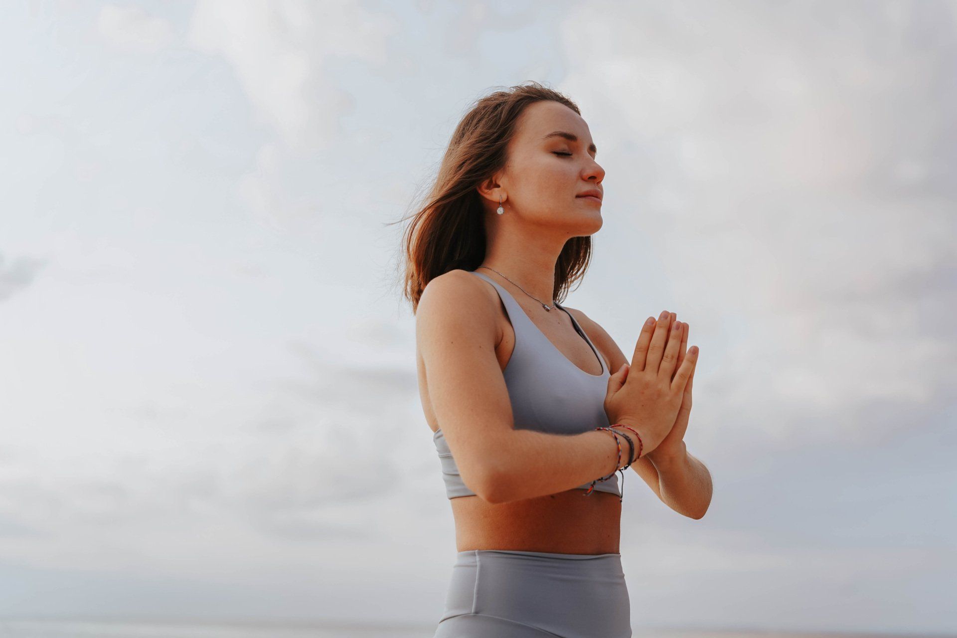 Woman in exercise clothes with hands in prayer, eyes closed, against a cloudy sky.