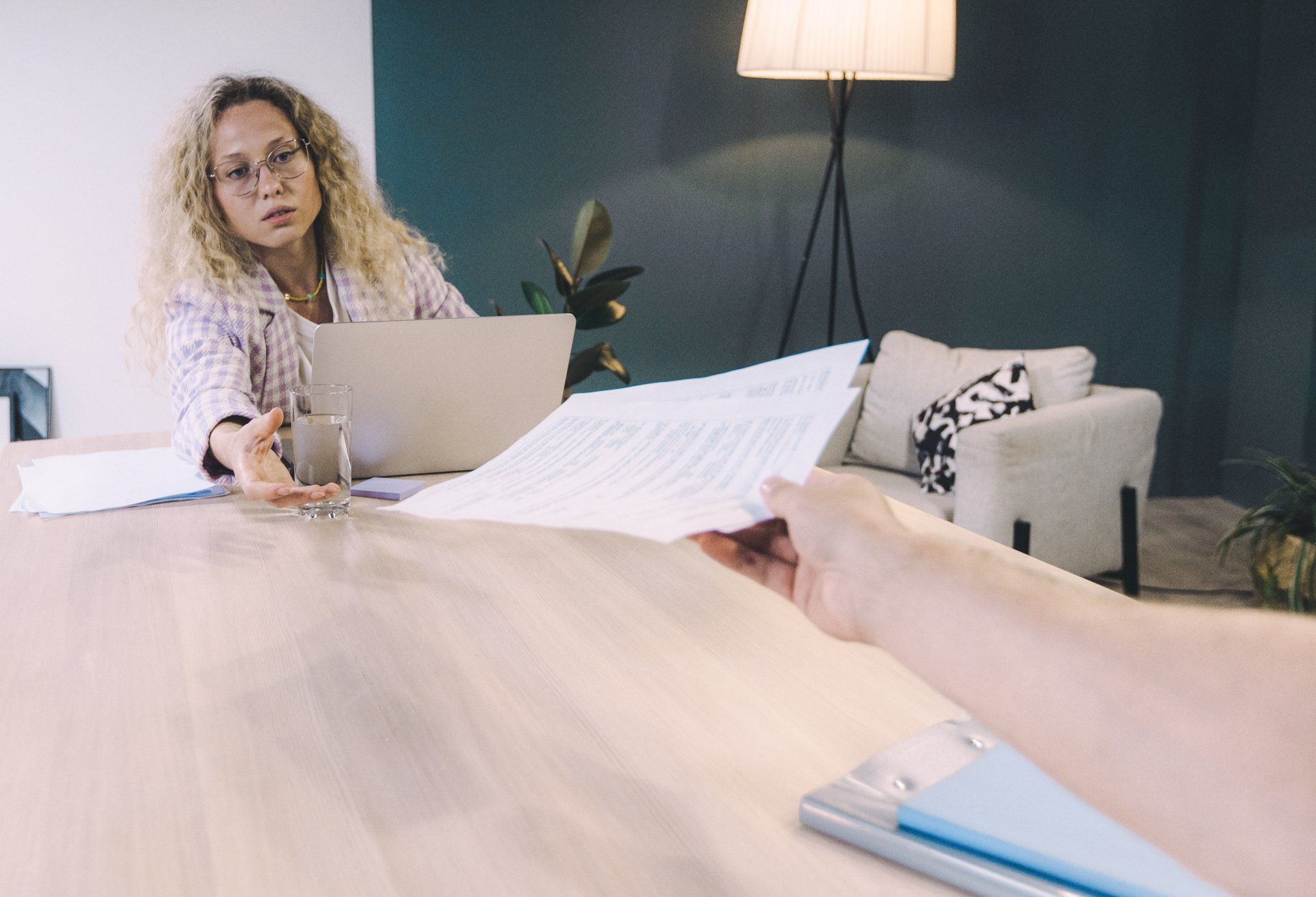 A woman is sitting at a table with a laptop and a person is holding a piece of paper.