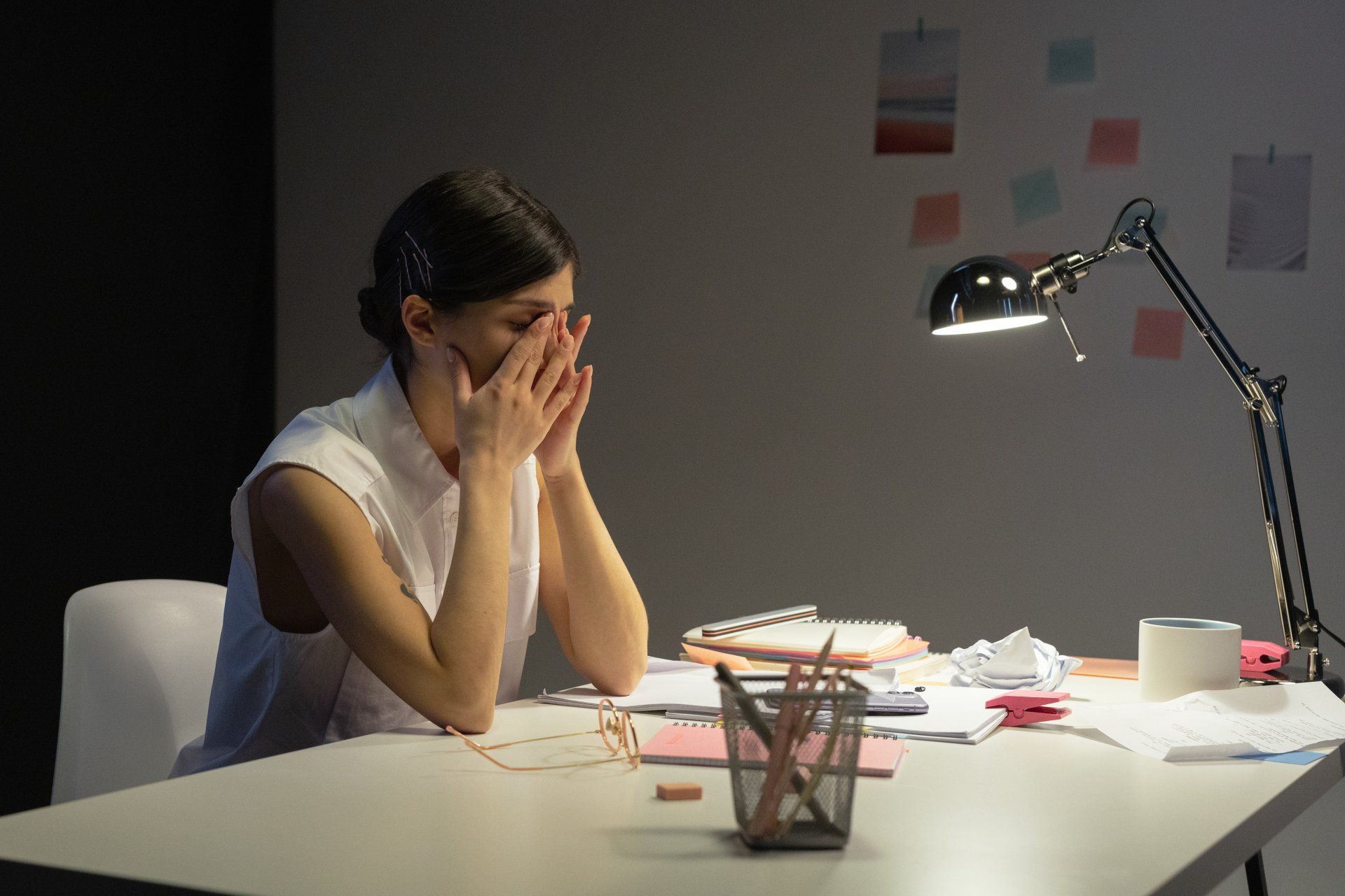 Woman with hands on face, looking stressed at a messy desk under a desk lamp.