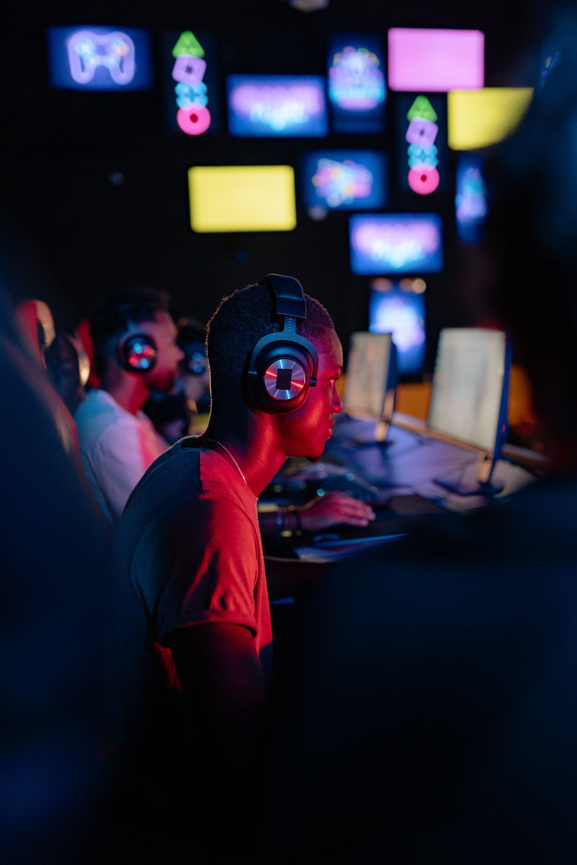 Gamers in a dimly lit room, illuminated by screens. Person with headphones focused on a computer. Red and blue lighting.