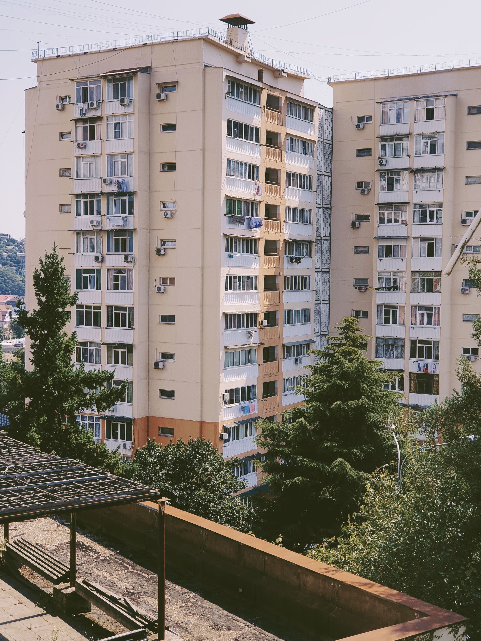 Two beige apartment towers with numerous windows and balconies set against a backdrop of trees and a clear sky.