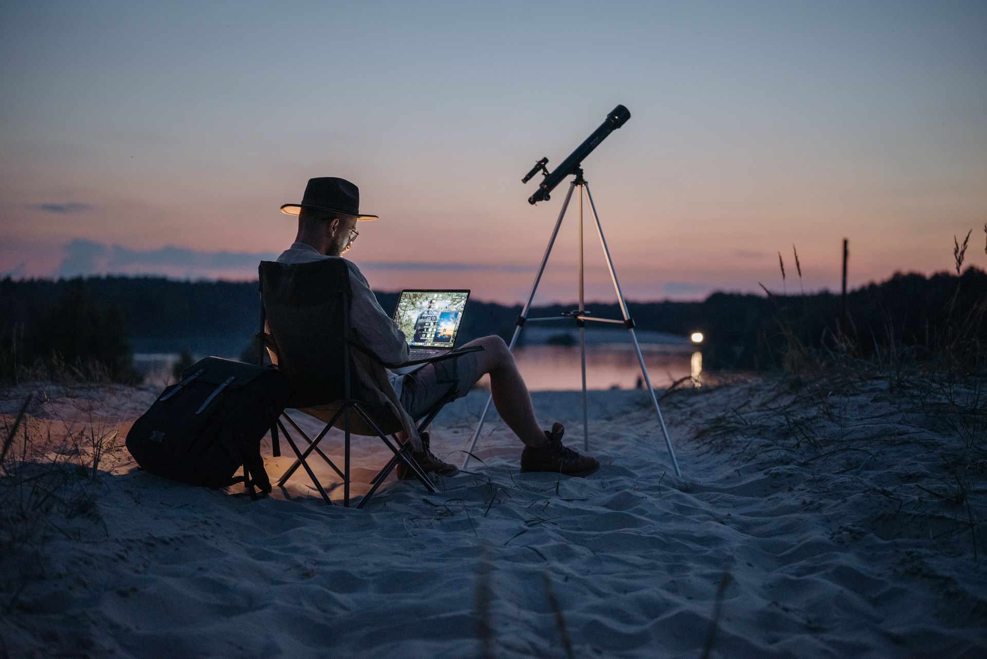 A man is sitting on a beach using a laptop and a telescope.