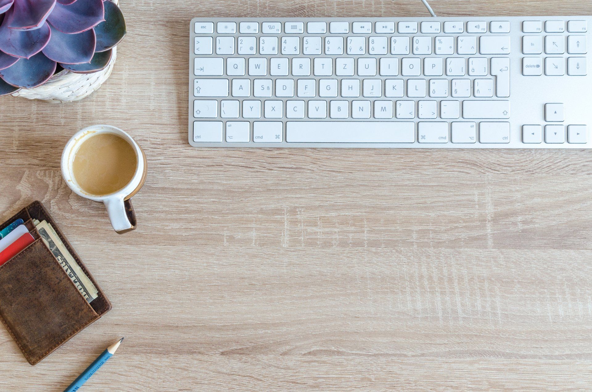 A wooden desk with a keyboard , a cup of coffee , a wallet , and a plant.