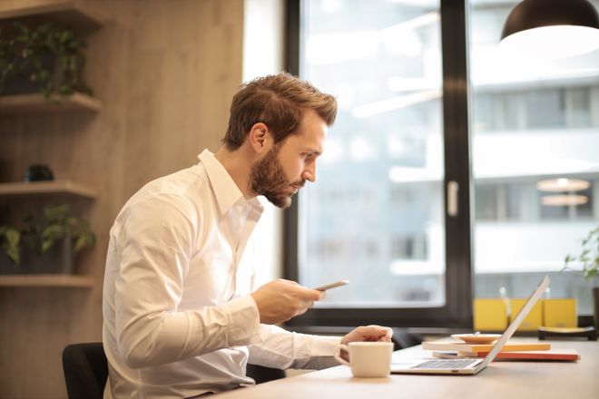 Man in a white shirt working at a laptop in a bright office, holding a cup and looking at a phone