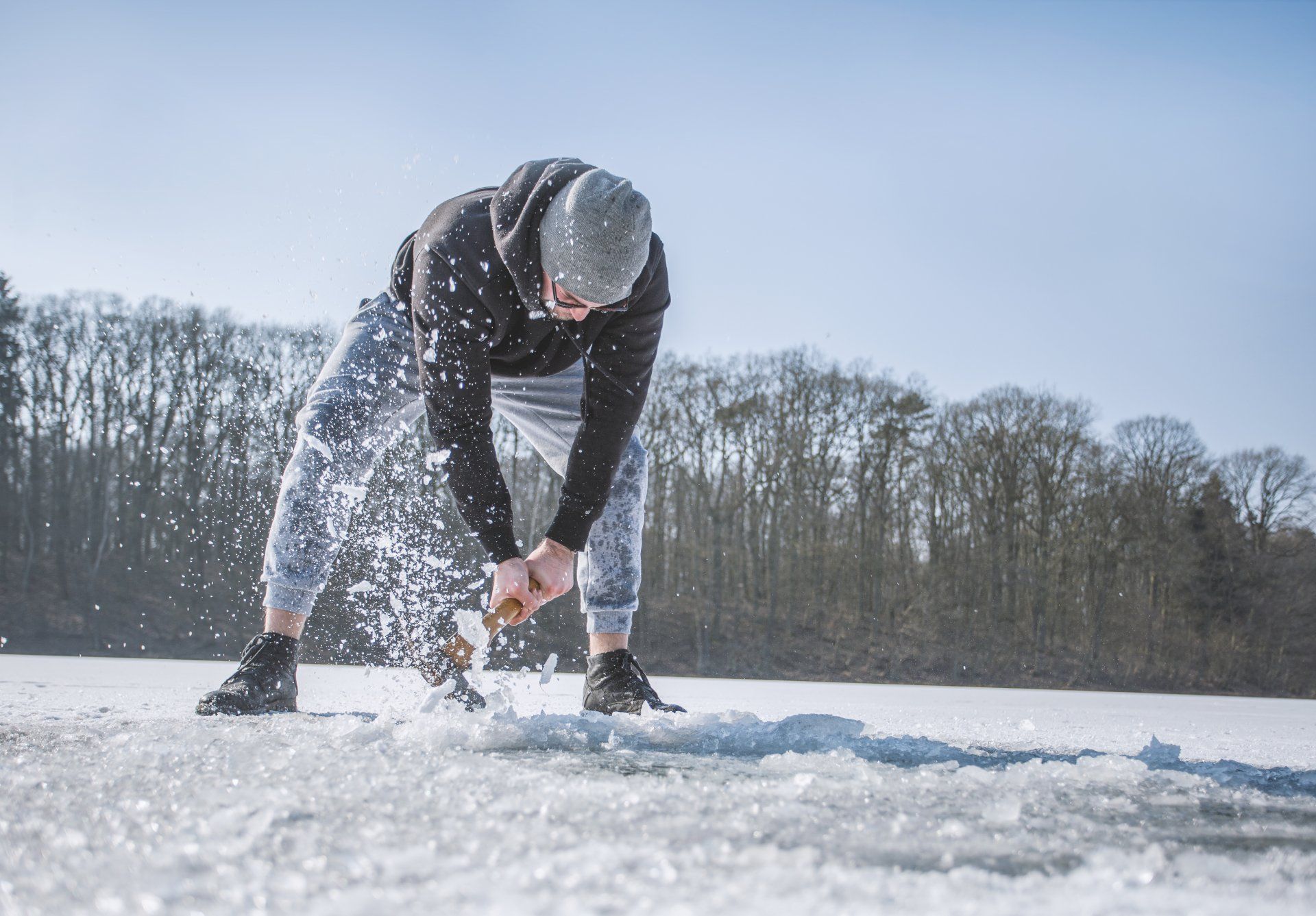 Eine Person bohrt Eis auf einem zugefrorenen, schneebedeckten See; im Hintergrund sind Bäume zu sehen.