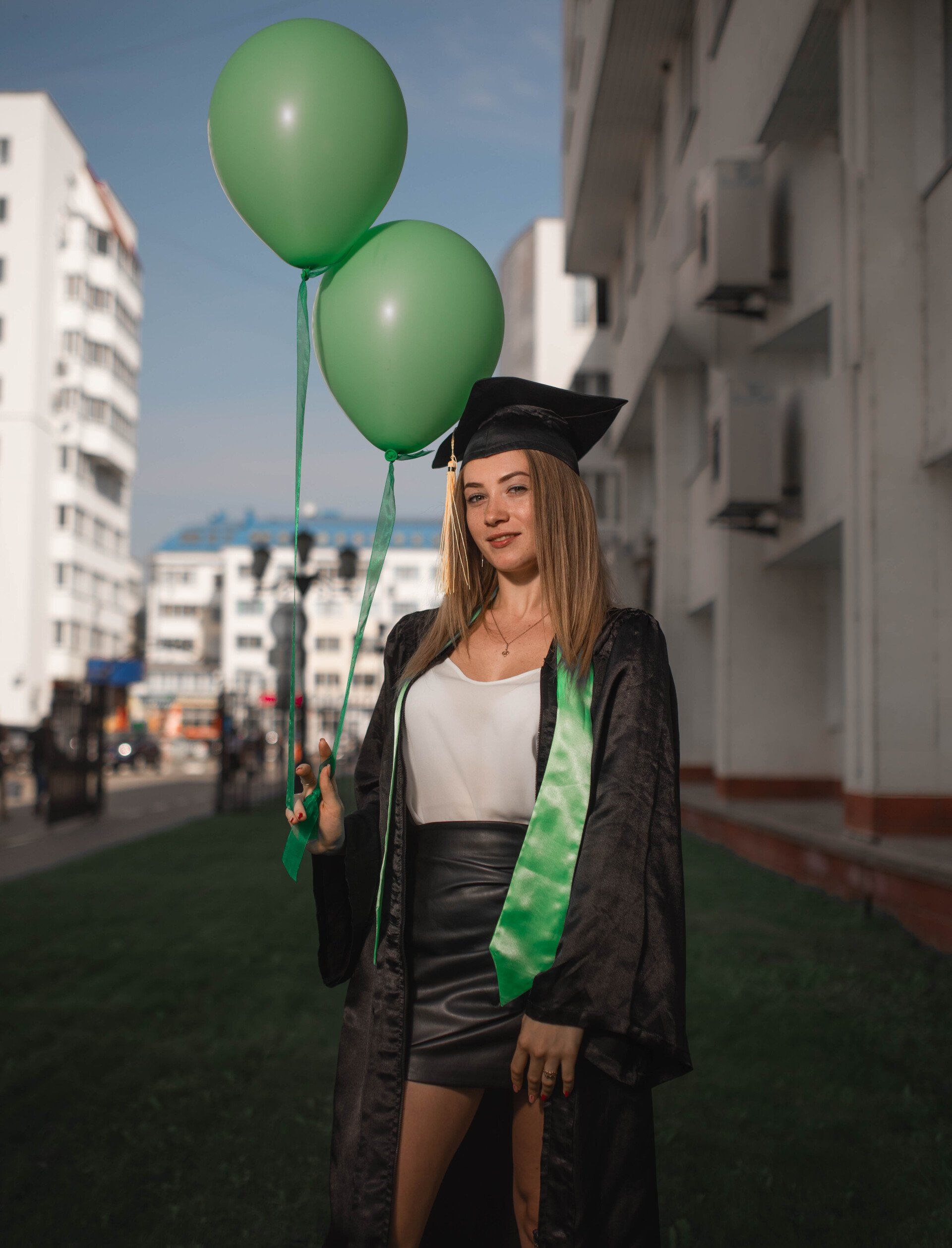 Graduating woman in cap and gown holds green balloons, smiles in front of a building.