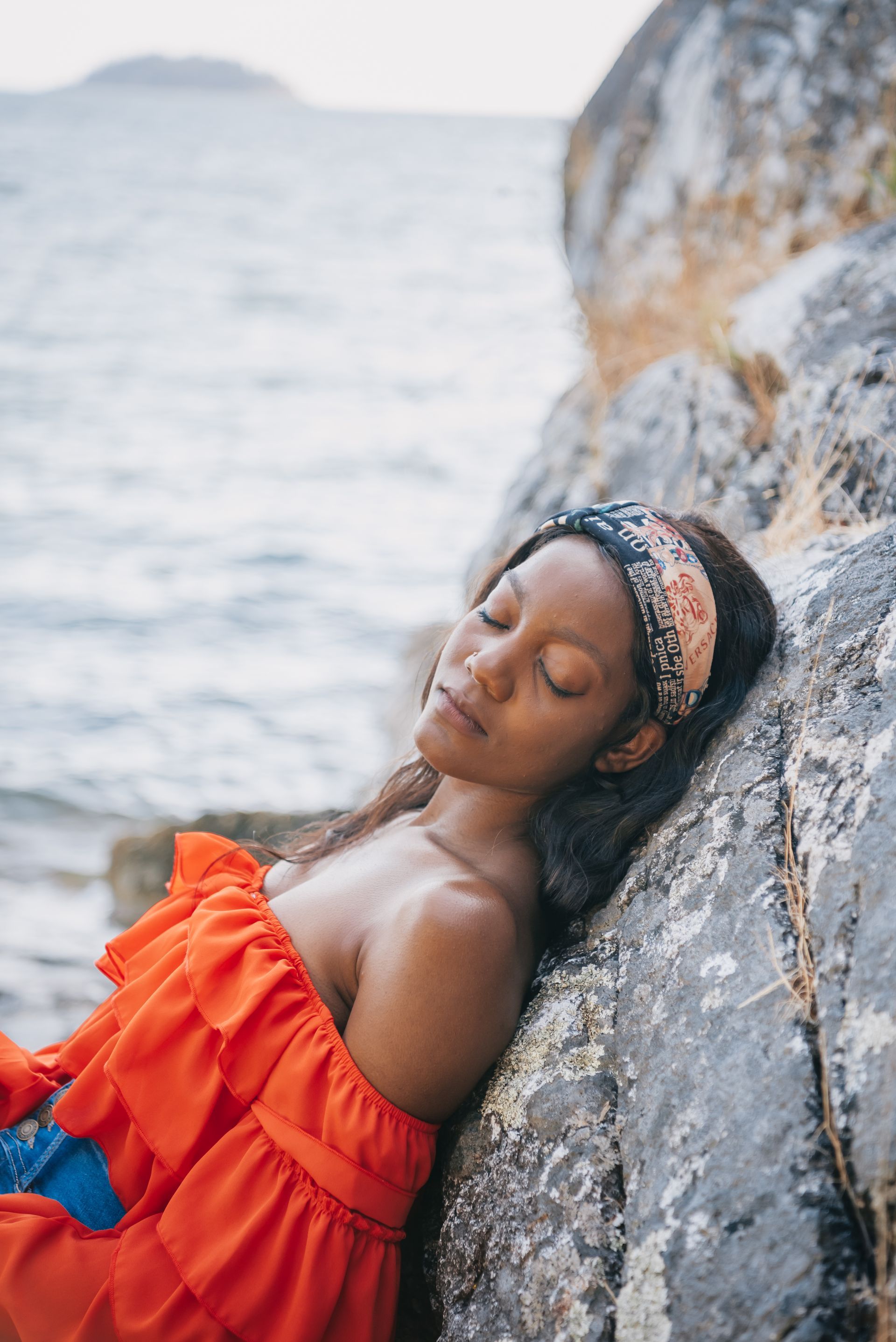 A woman in an orange dress is laying on a rock near the water.