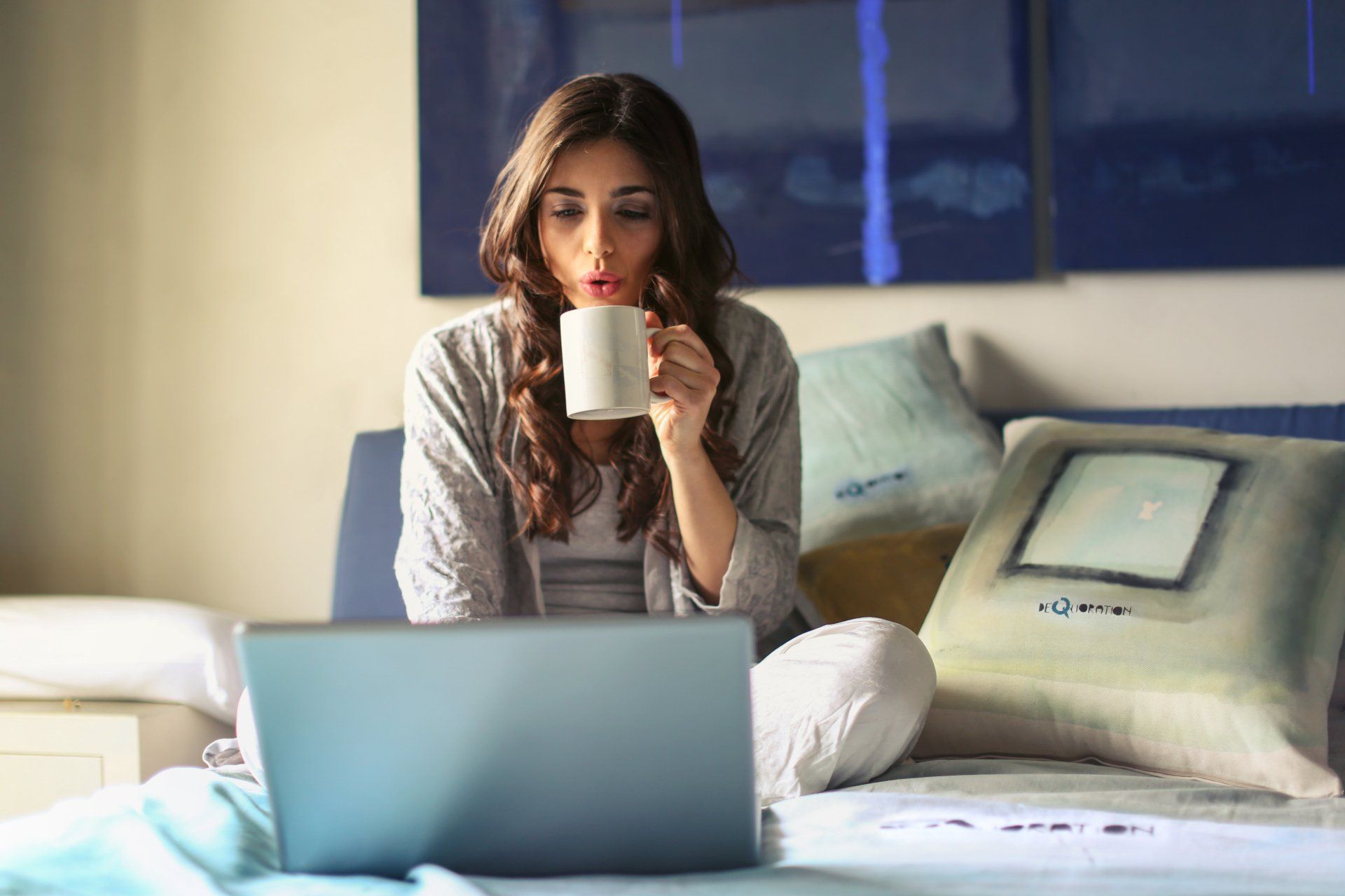 Woman sitting on bed, drinking from a mug while looking at a laptop.