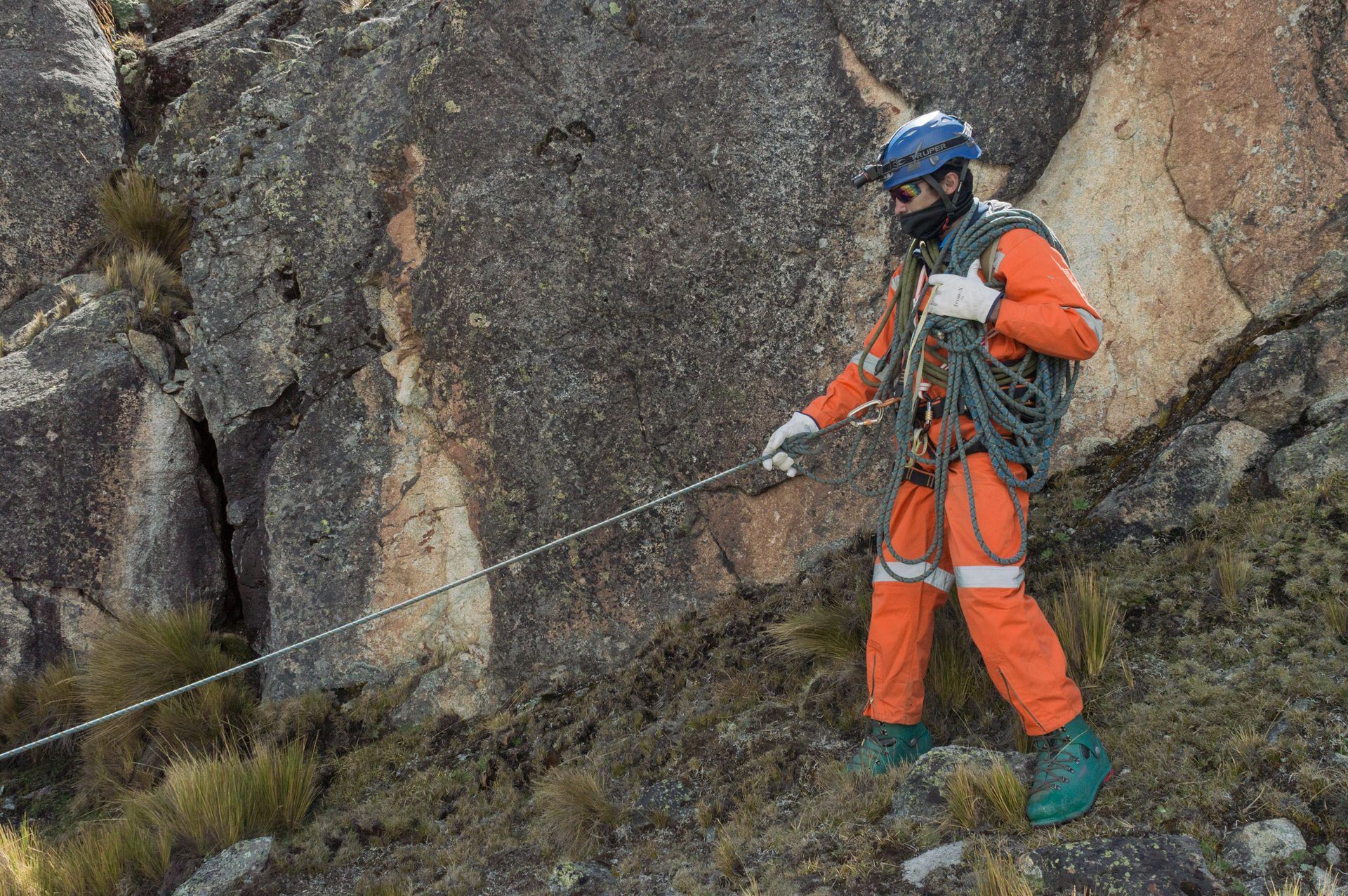 Person in orange gear on rocky terrain, pulling cable, safety helmet and gloves.