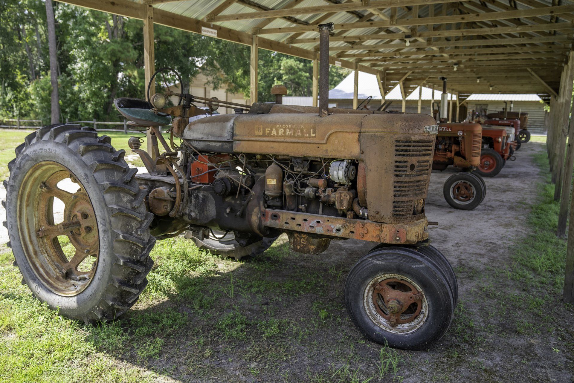 A row of old rusty tractors are parked under a shed.