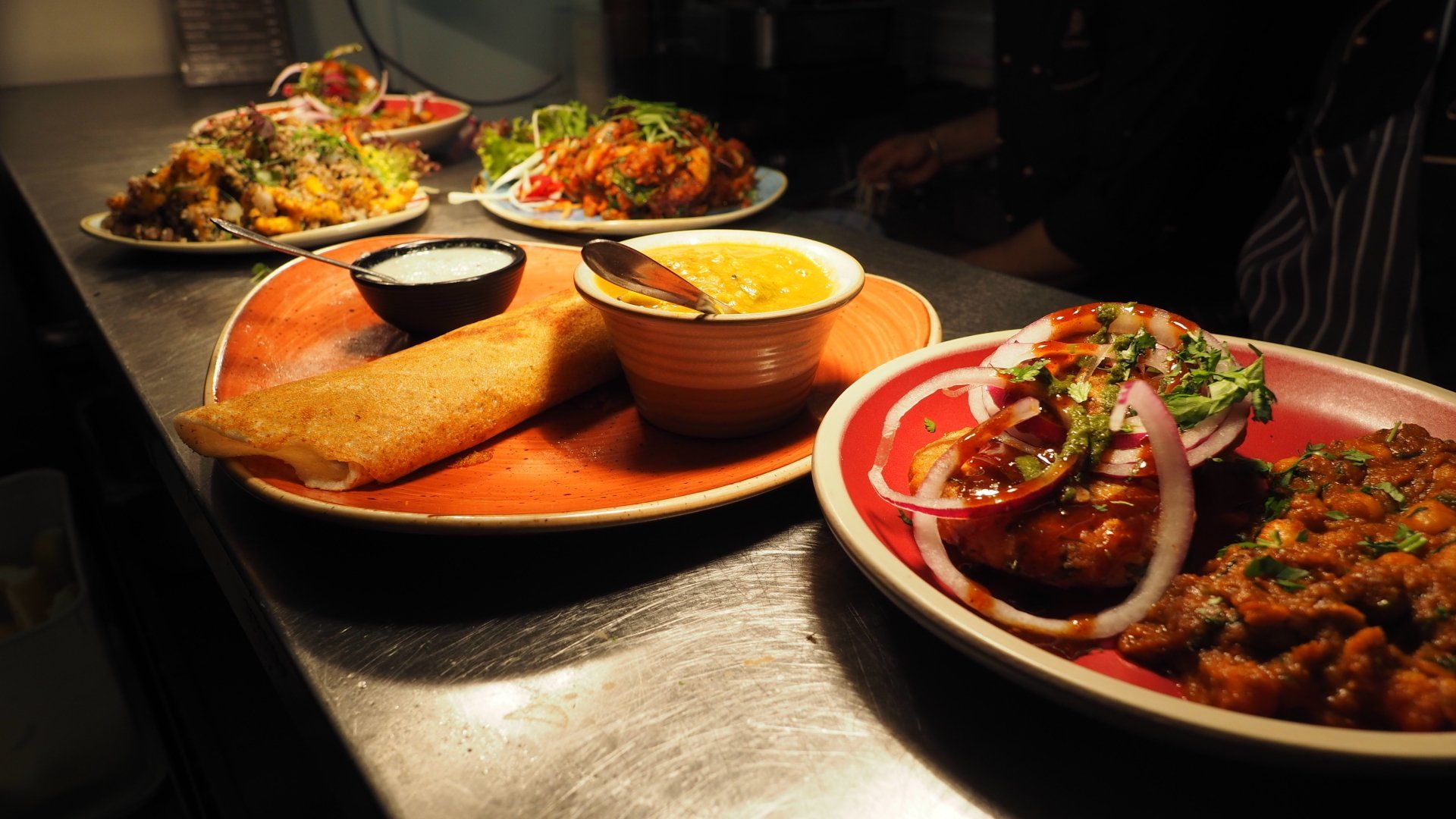 Plates of Indian food on a counter, including curry, rice, and a wrap, with a server in the background.