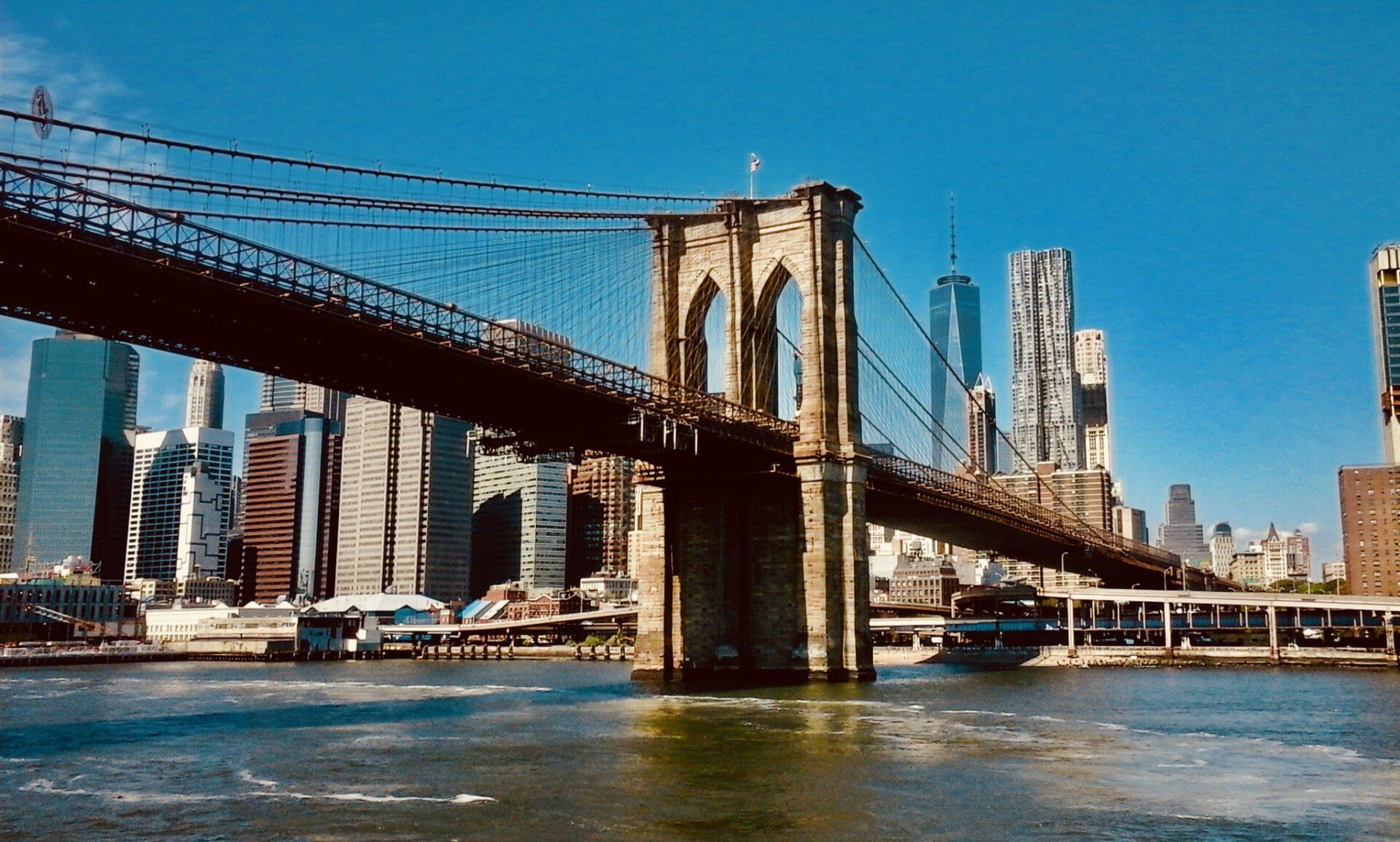 Brooklyn Bridge spanning the East River, with Manhattan skyline in background on a sunny day.