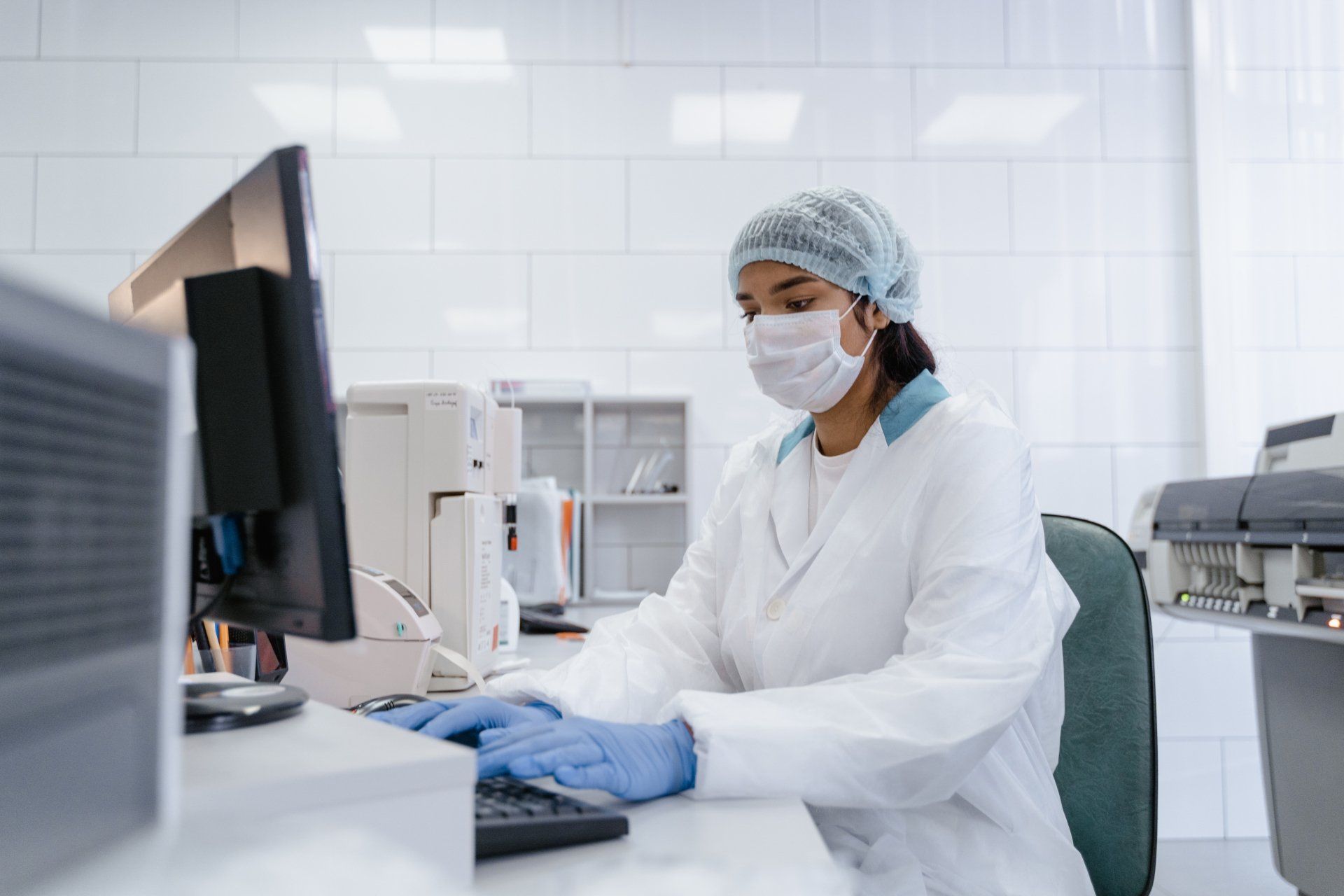 A female scientist wearing a mask and gloves is using a computer in a laboratory.