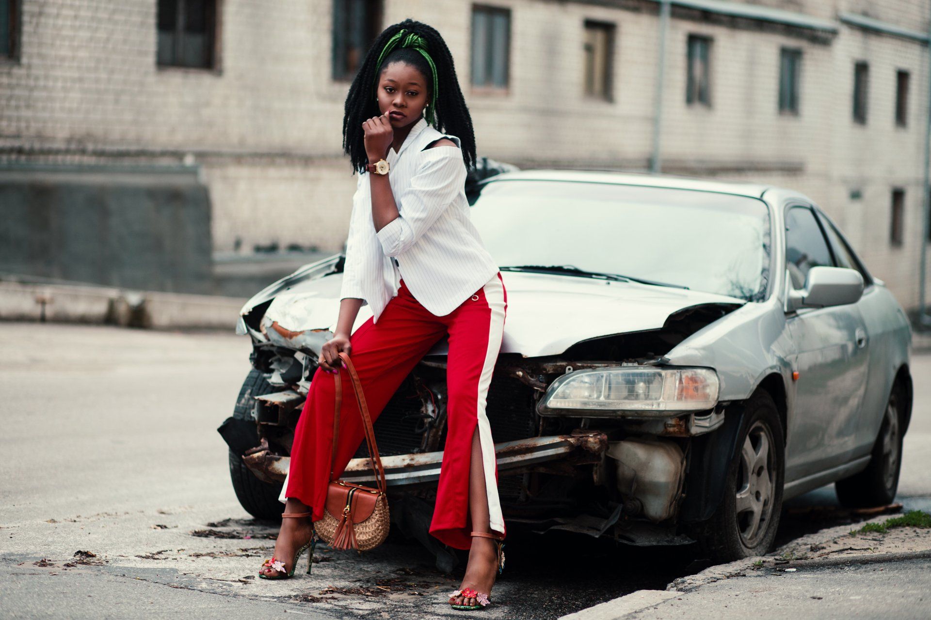 Woman in red pants leaning against a damaged black car on a city street
