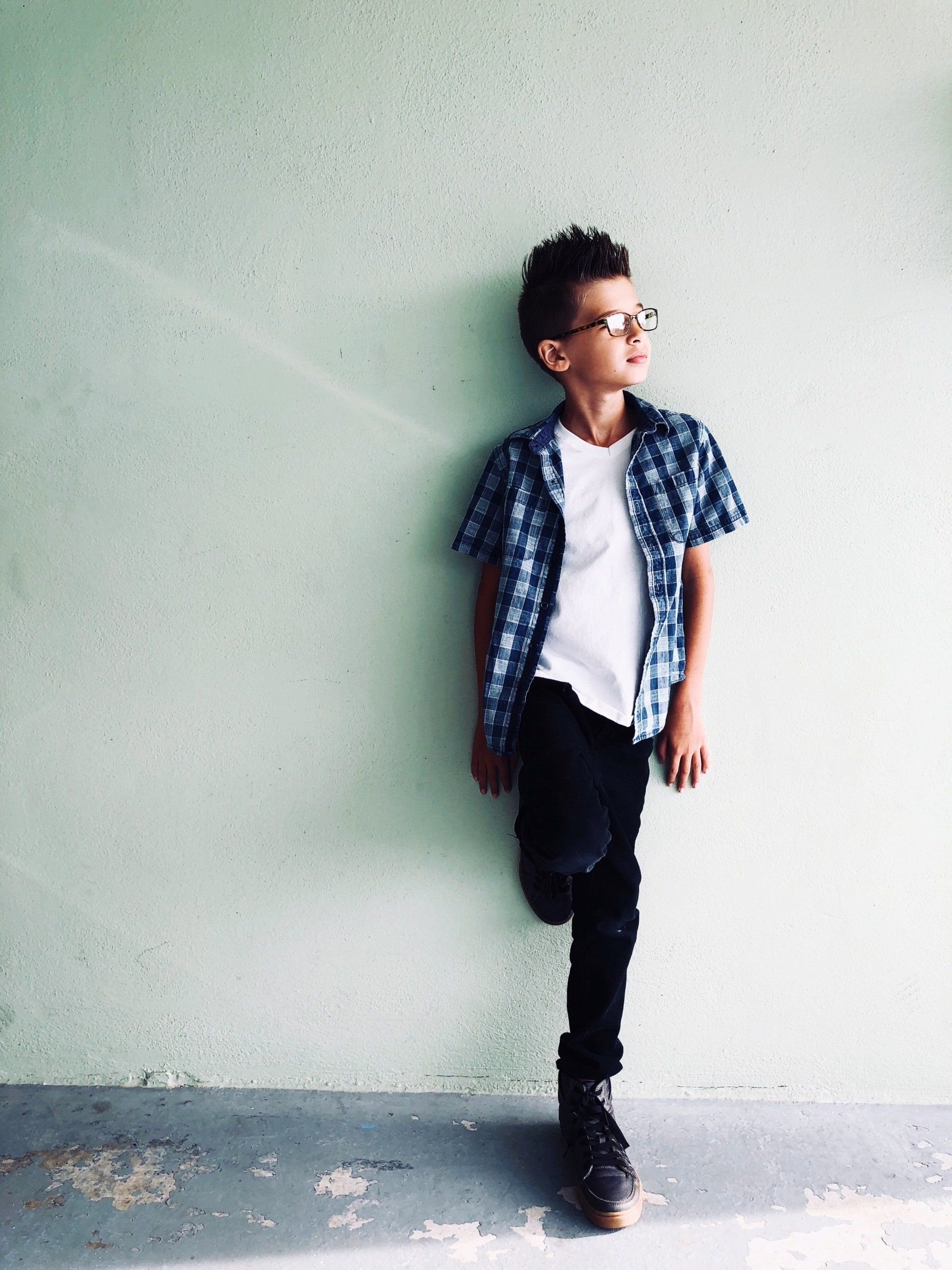 Boy with glasses leaning against a light green wall, wearing blue plaid shirt, white tee, black jeans, and boots.