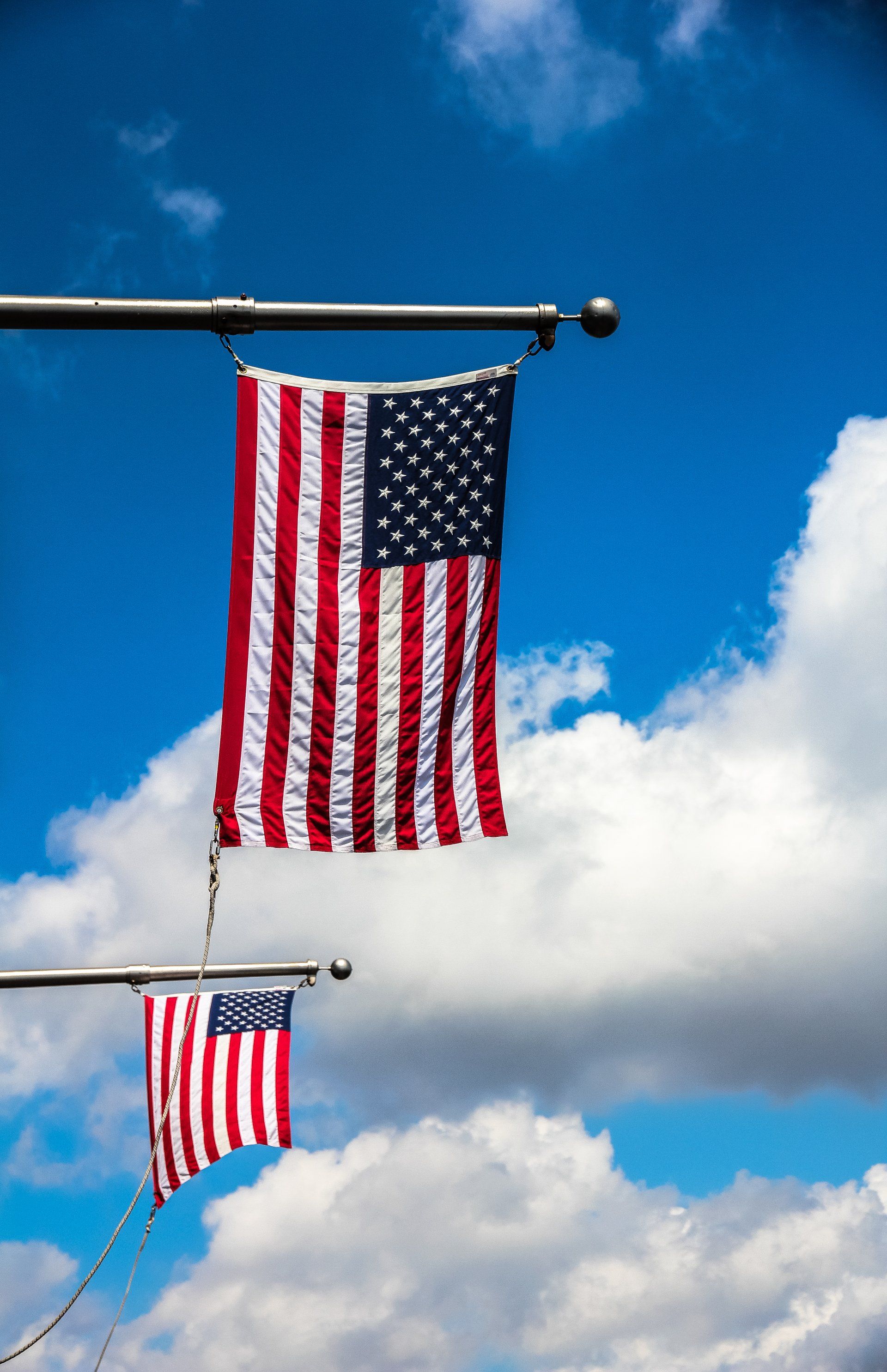 Two American flags hanging against a bright blue sky with fluffy white clouds.