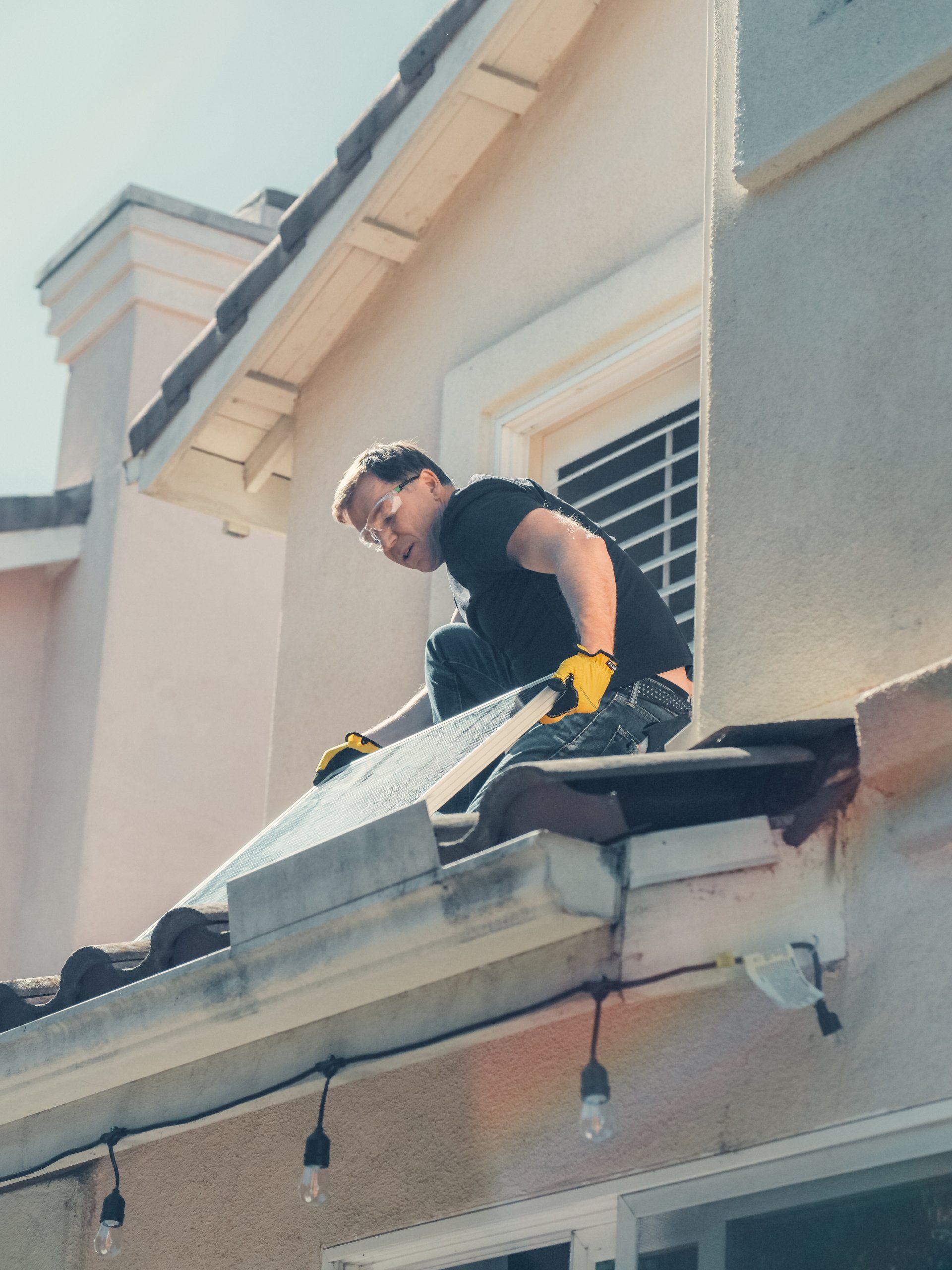 Worker in black shirt and yellow gloves on a roof, inspecting the edge of a house.