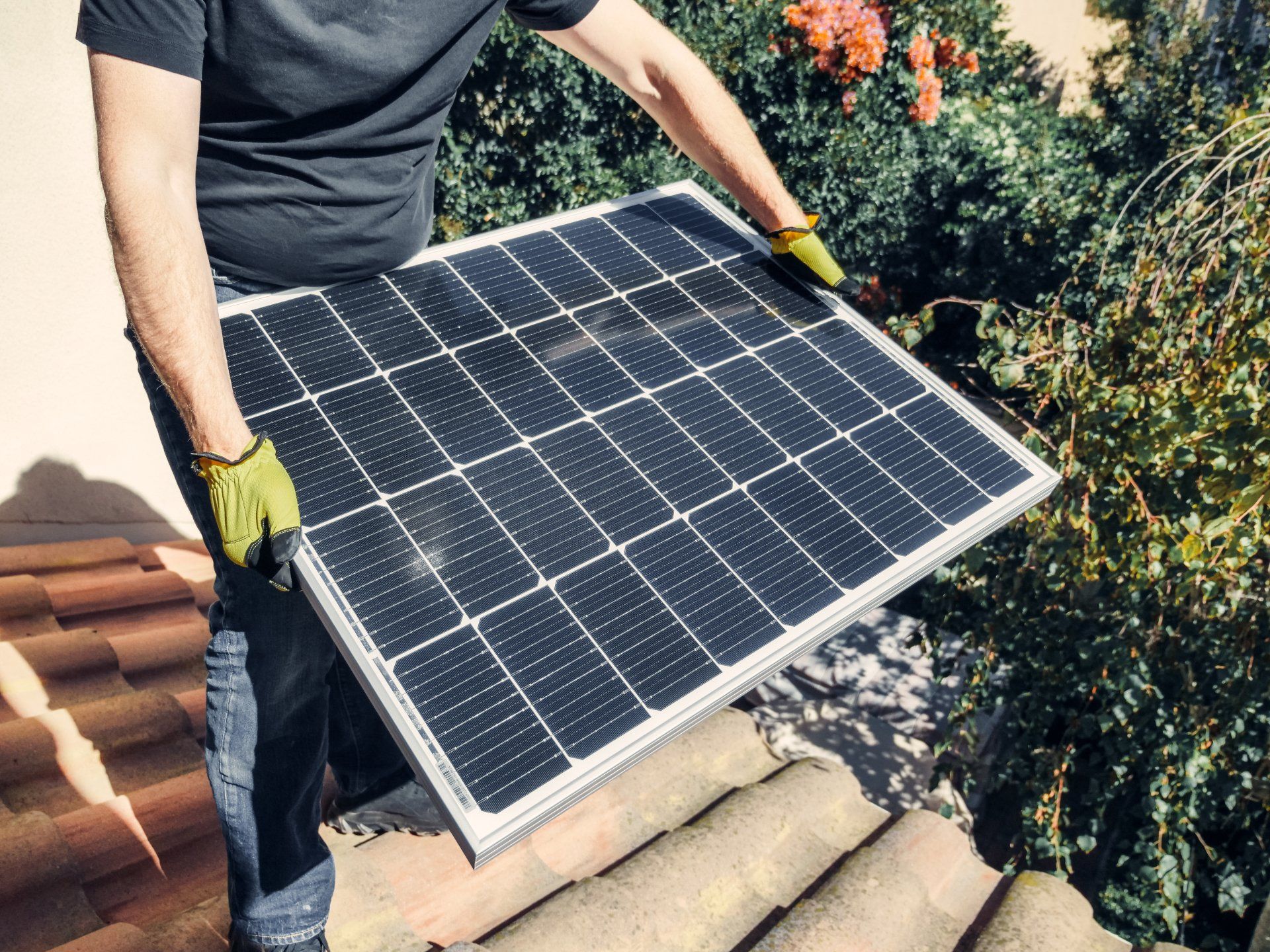 Person carrying a solar panel on a rooftop beside trees and terracotta tiles