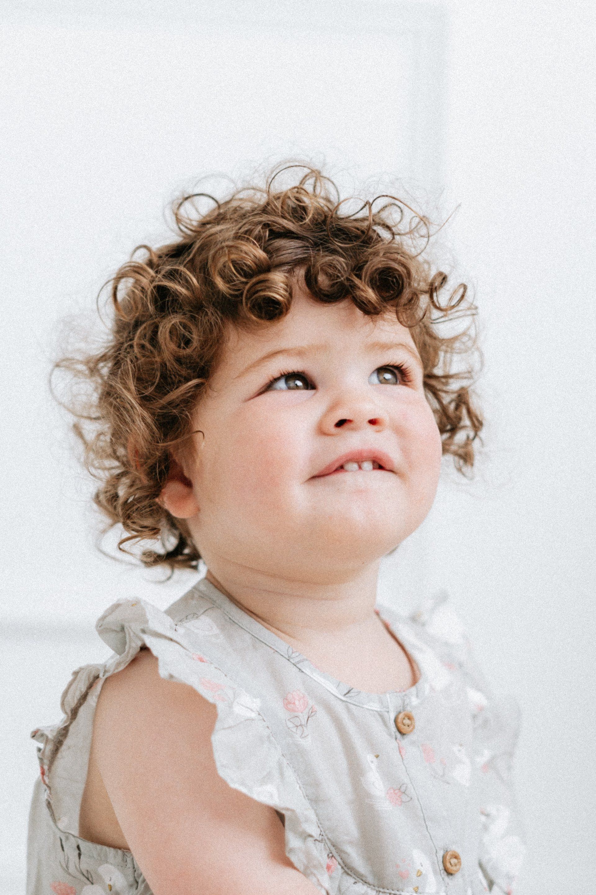 Curly-haired toddler looking upward with a slight smile, wearing a light-colored dress against a white background.