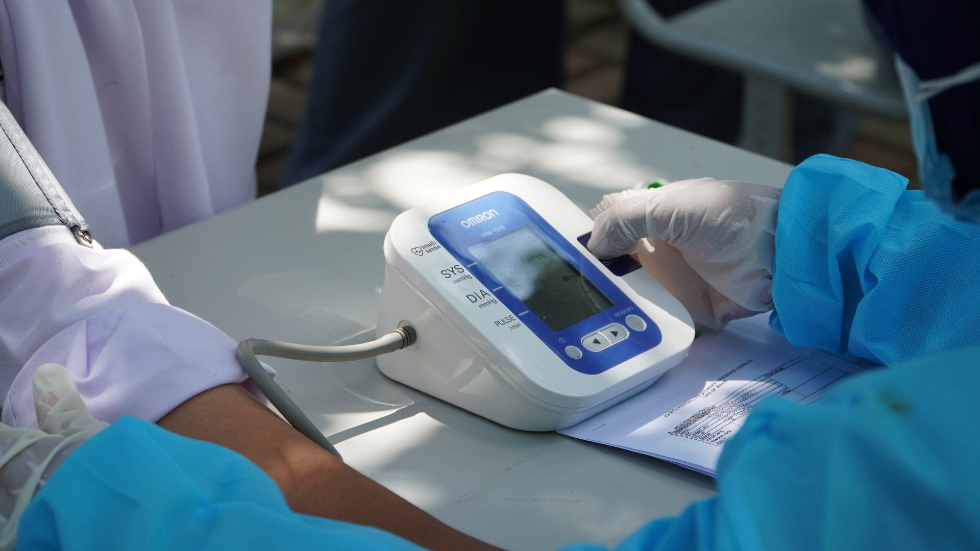 Person's arm has blood pressure checked with a digital monitor; health worker in blue scrubs, gloves.