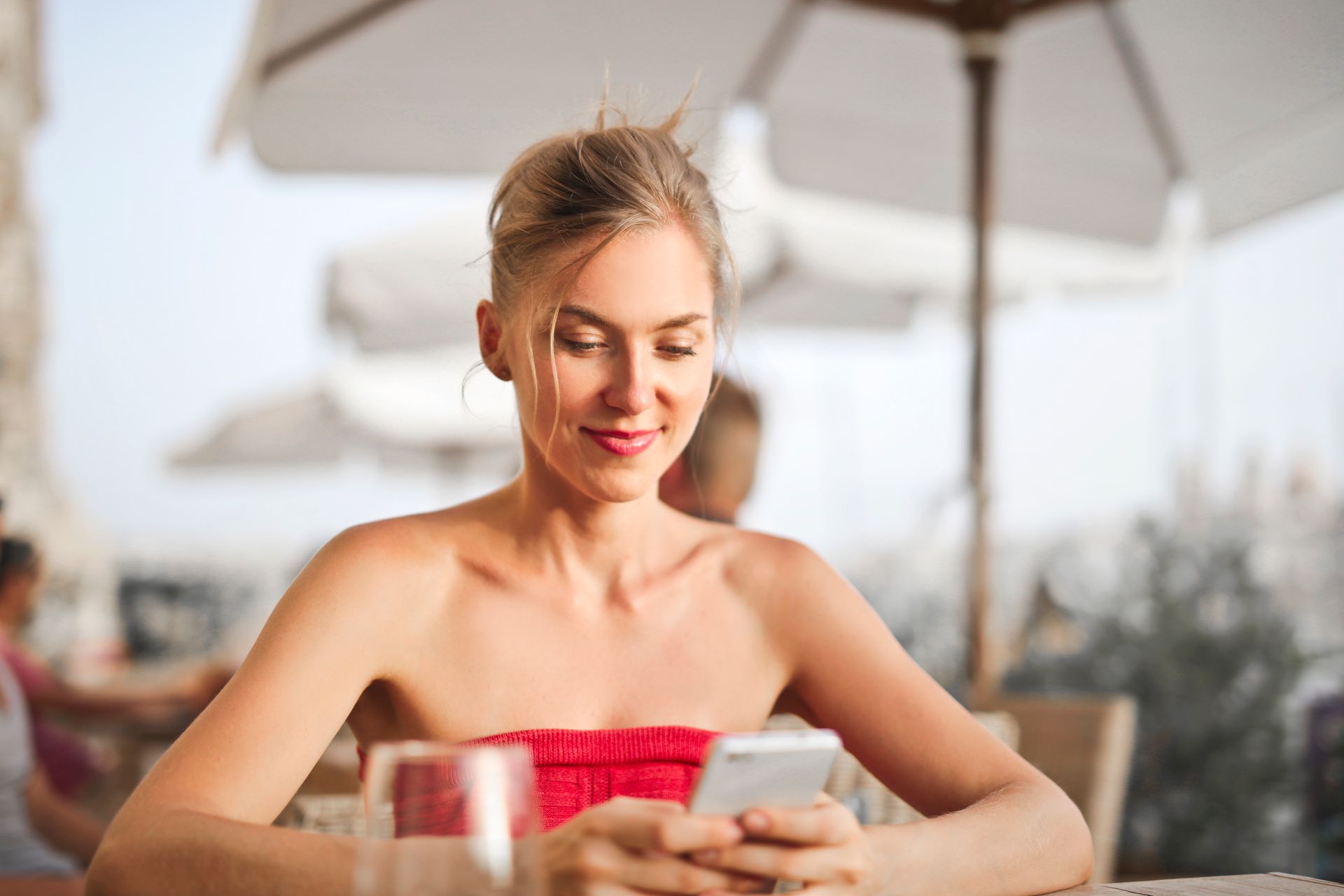 Blonde woman in red top, smiling, looking at phone at outdoor cafe table.