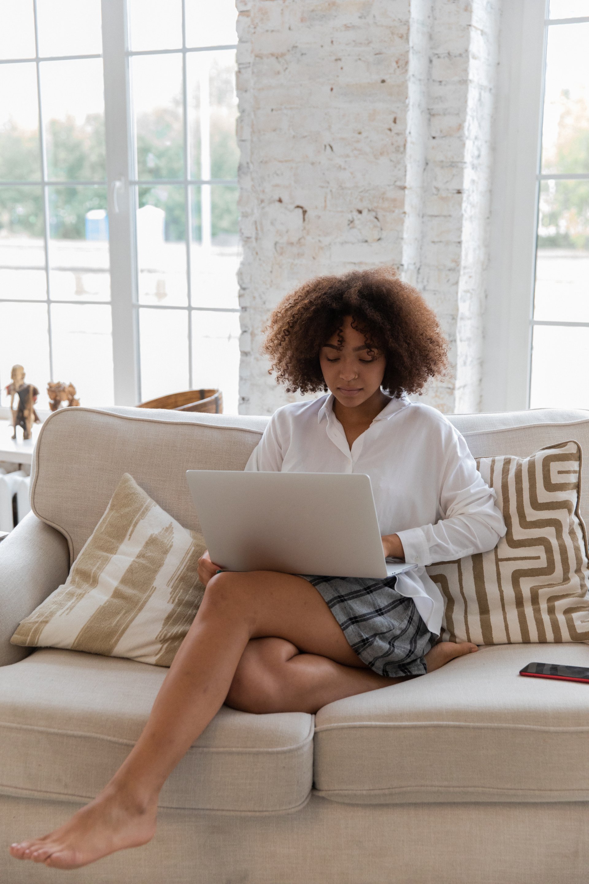 Woman with curly hair using laptop, sitting cross-legged on a beige sofa near a large window.