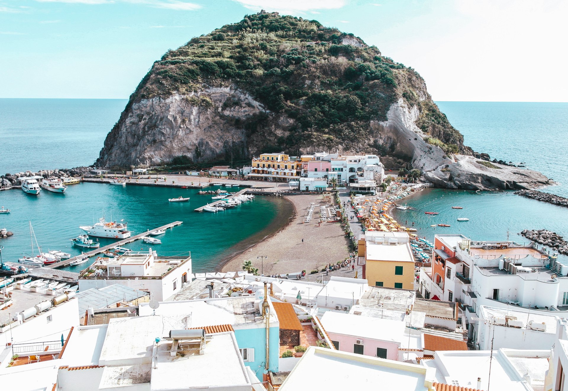 A coastal town with white buildings and a harbor, connected to a rocky, vegetation-covered island by a narrow beach.