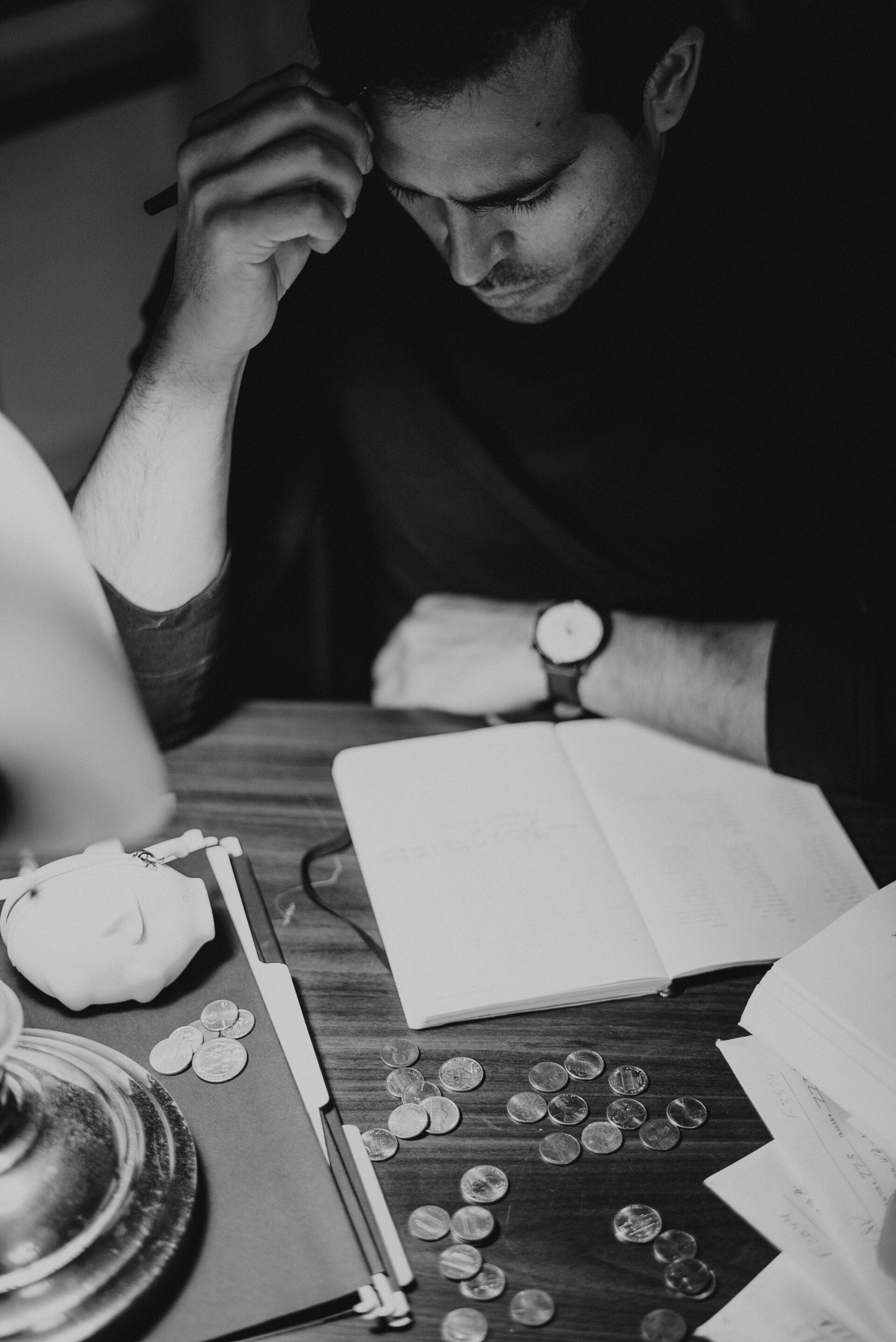 Man with pen, looking down at notebook, coins and papers on table. Black and white.