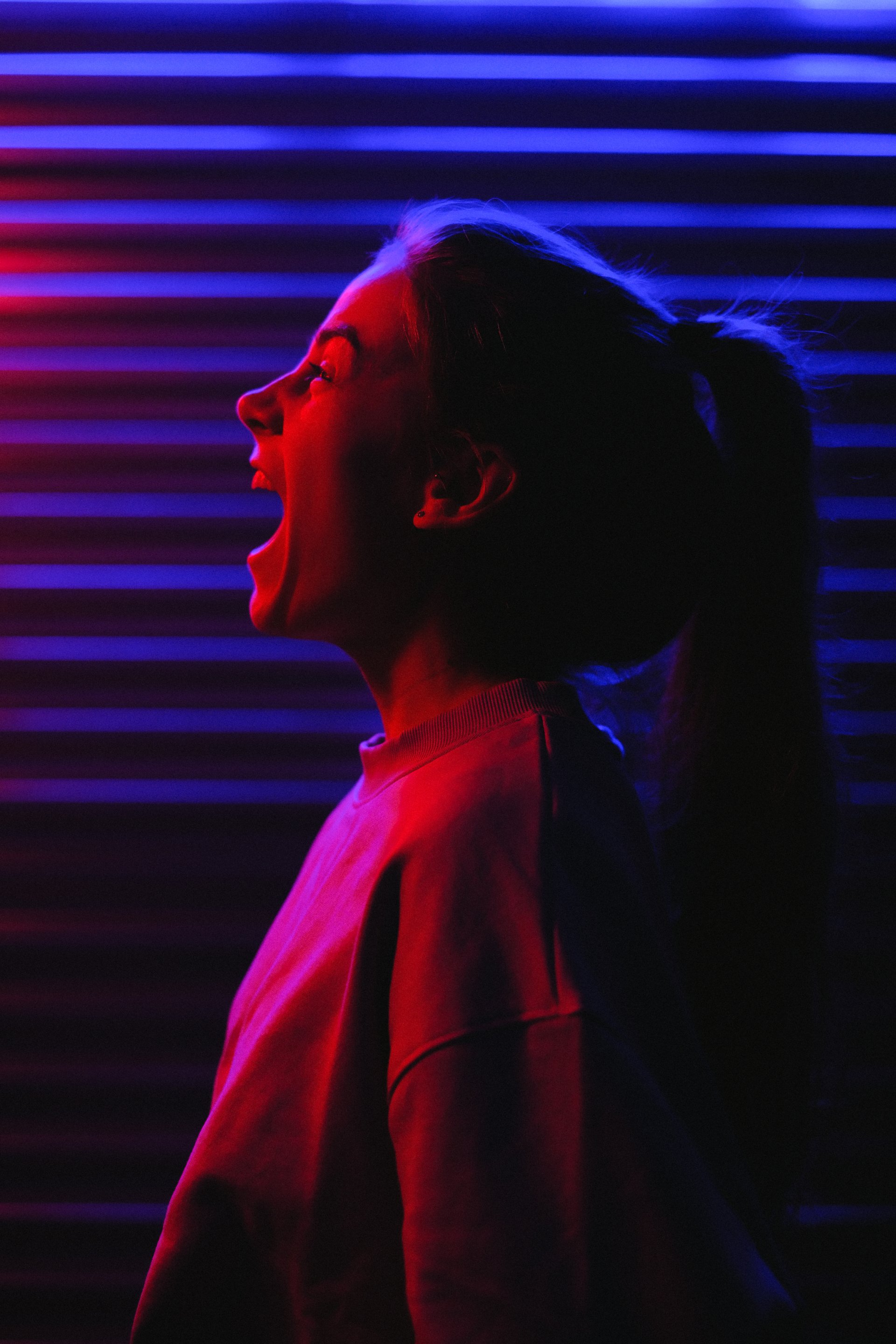 Woman silhouetted, mouth open in scream, lit by red and blue neon lights against a striped wall.