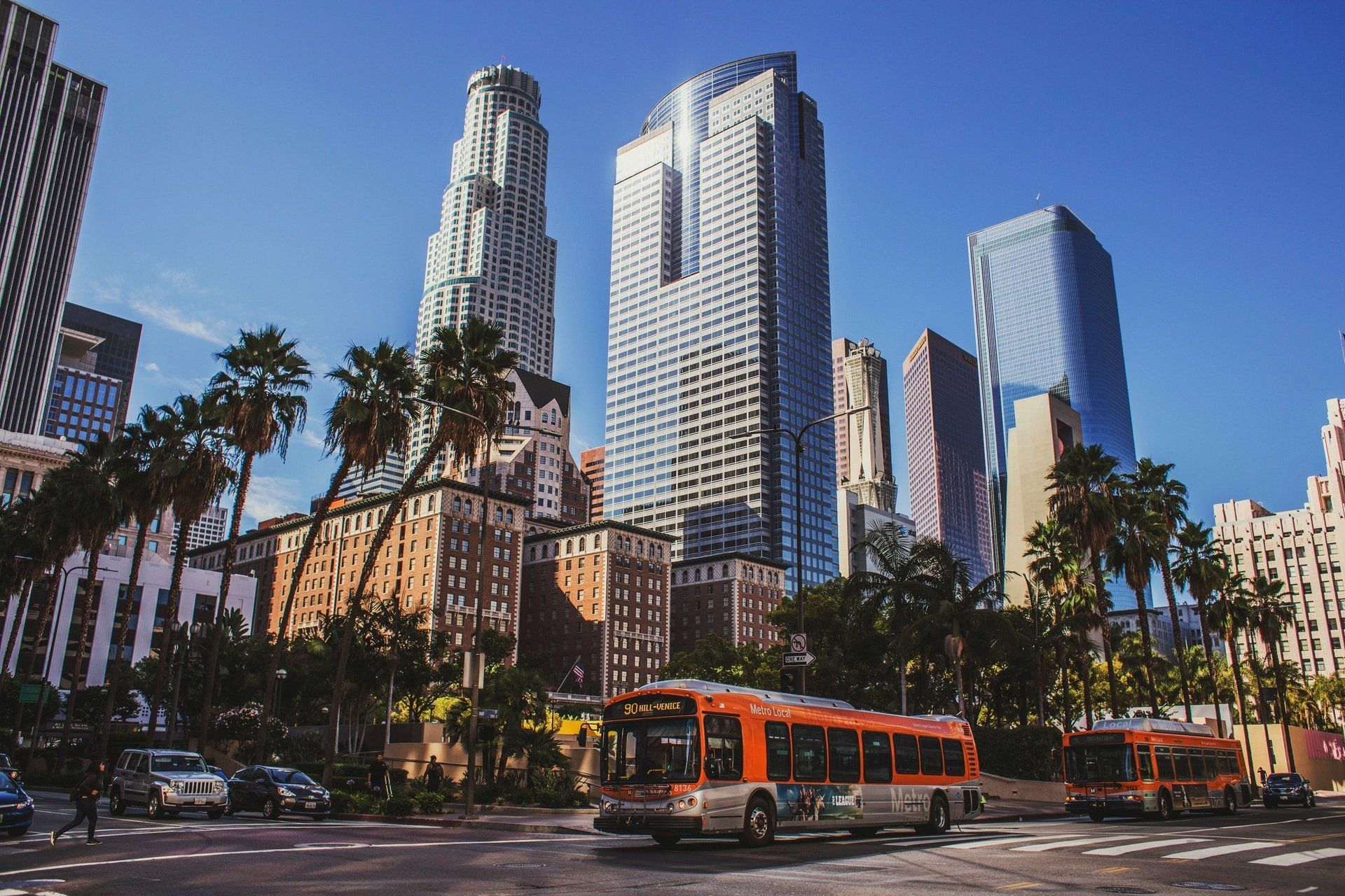 Downtown skyline with tall glass towers, palm trees, and orange city buses on a sunny street