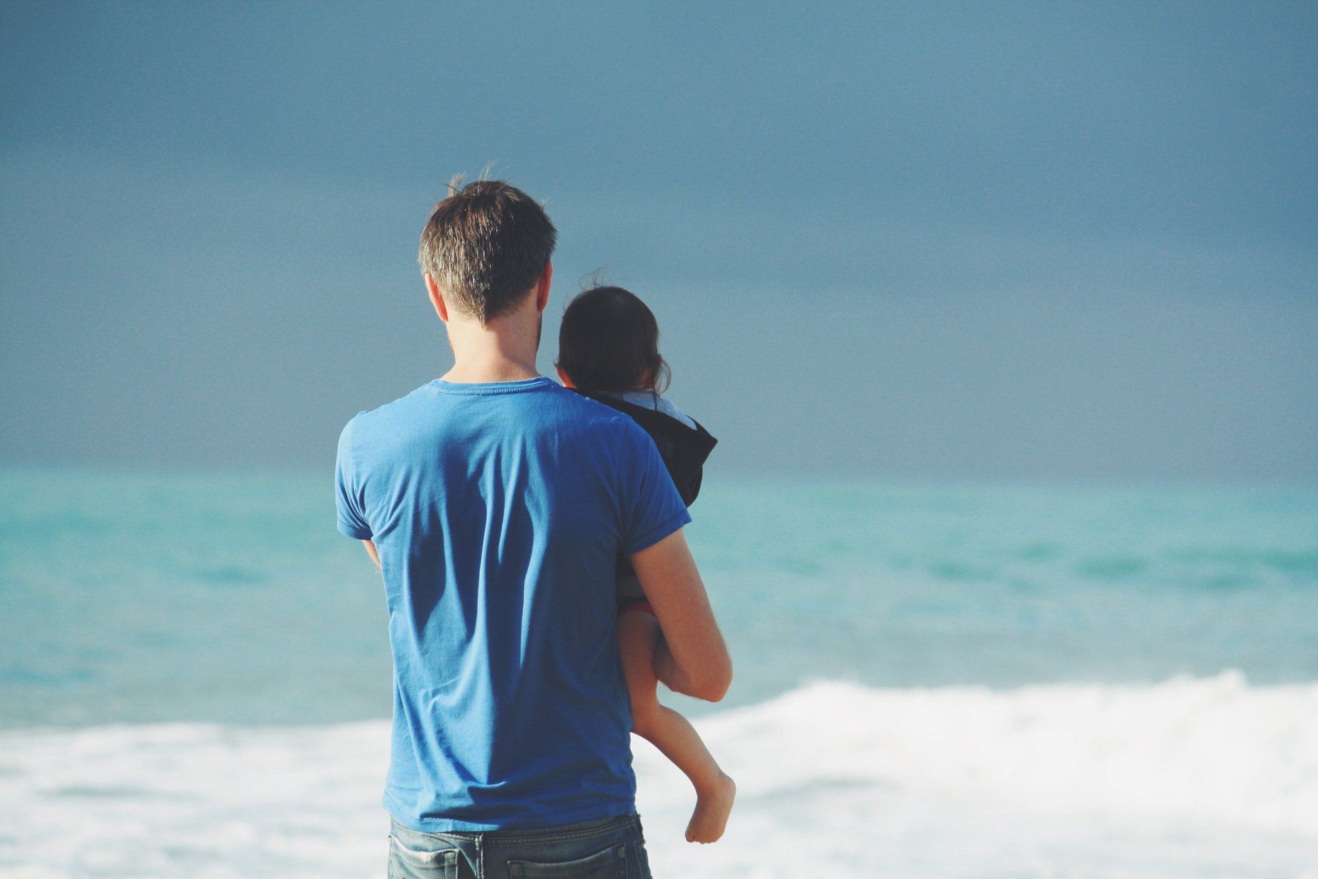 Man holding child, facing ocean under a cloudy sky.