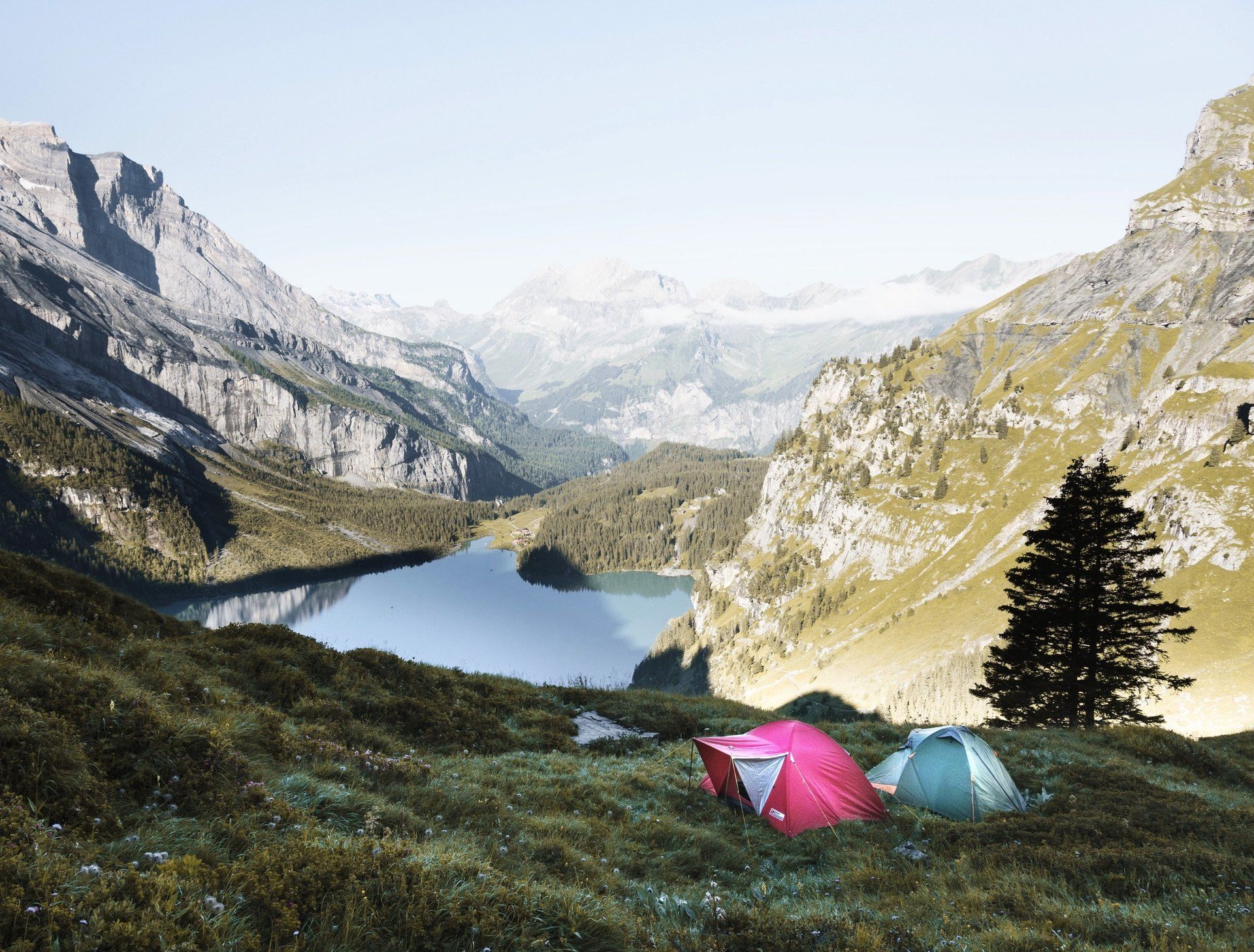Two tents are sitting on top of a grassy hill next to a lake in the mountains.