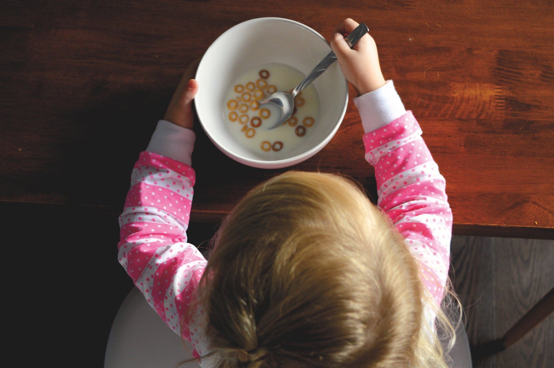 Child eating cereal from a white bowl at a wooden table. Pink striped sleeves.