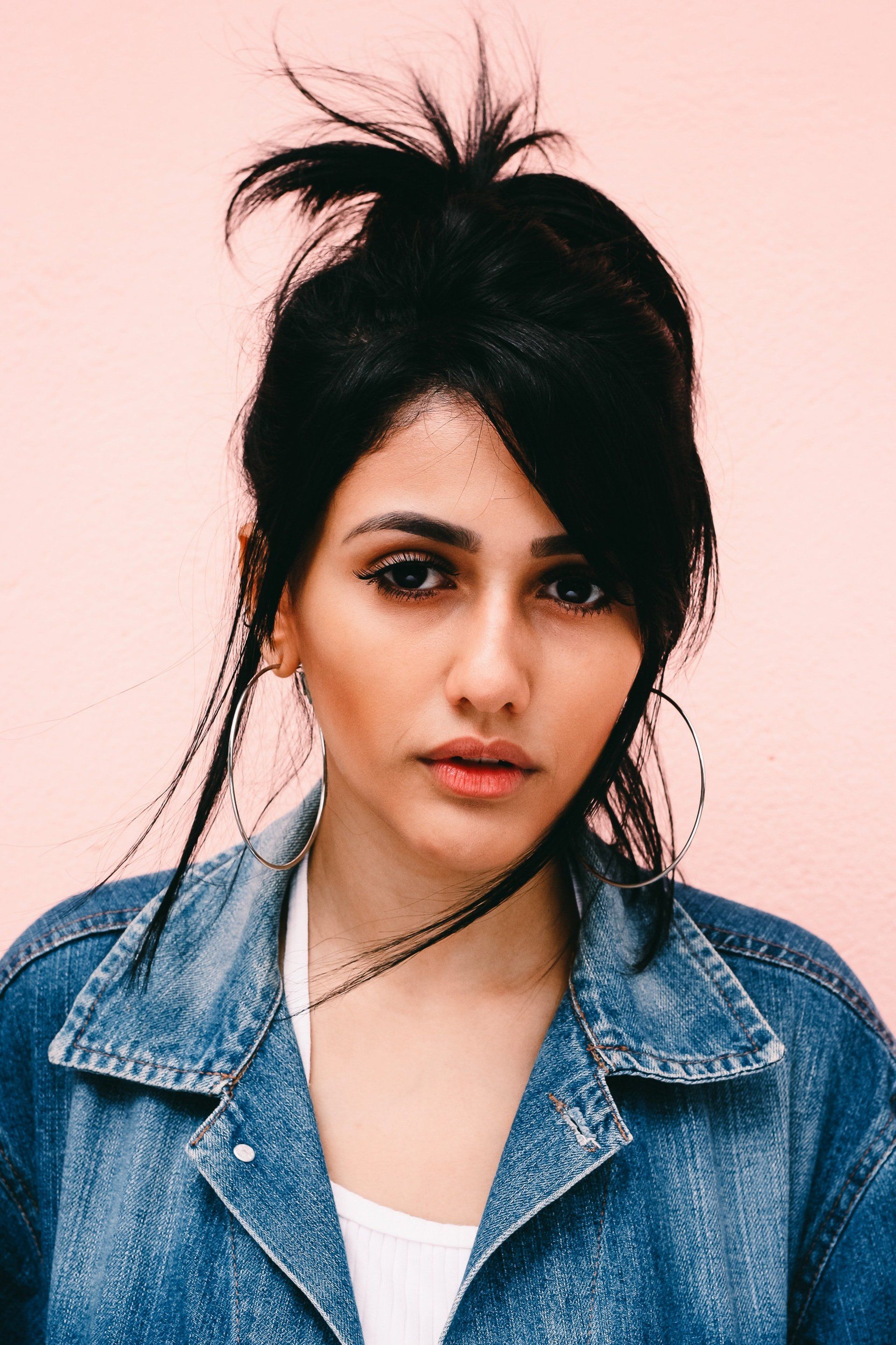 A woman wearing a denim jacket and earrings is standing in front of a pink wall.