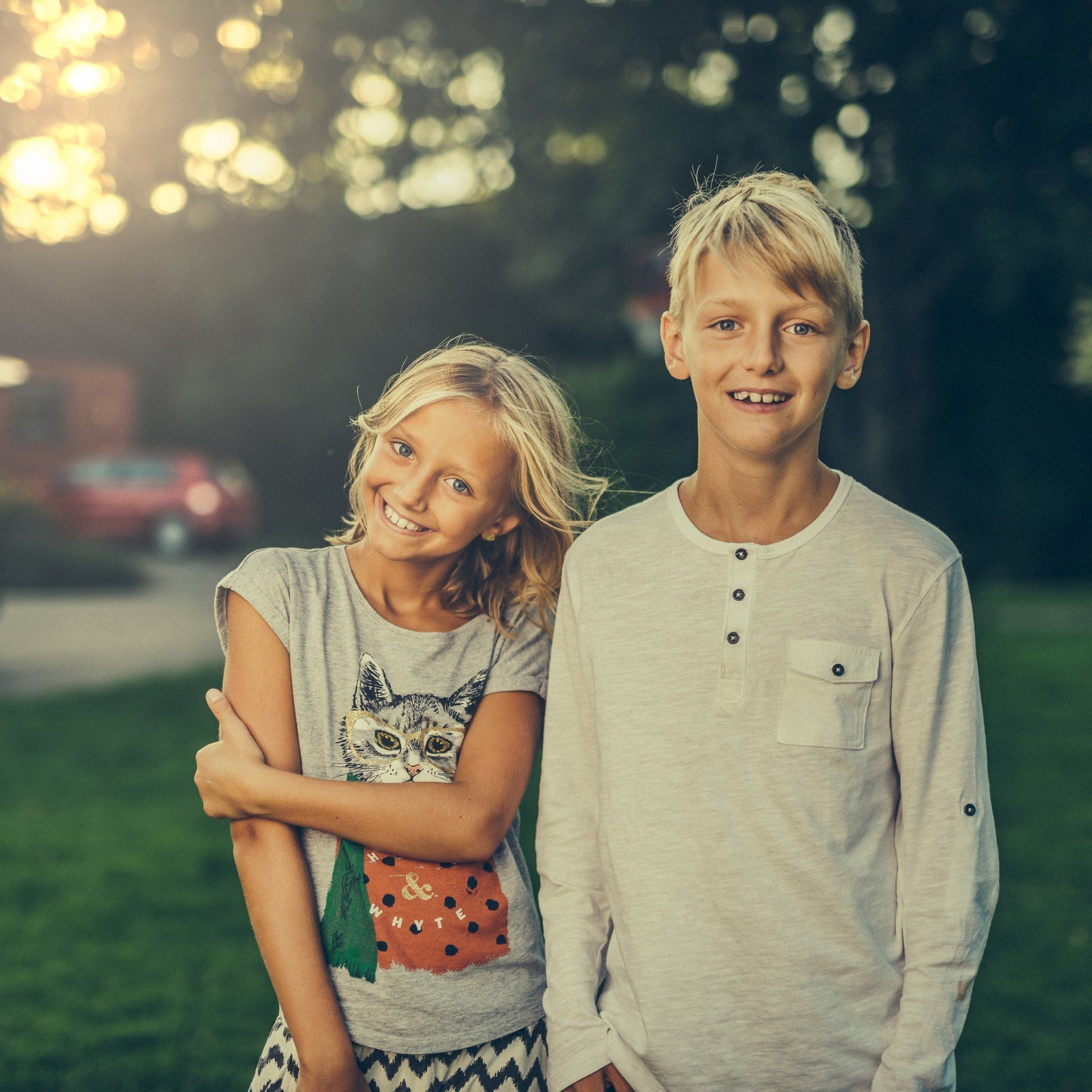 Two smiling children standing outdoors at dusk, one in a white shirt and the other in a light top and jeans.