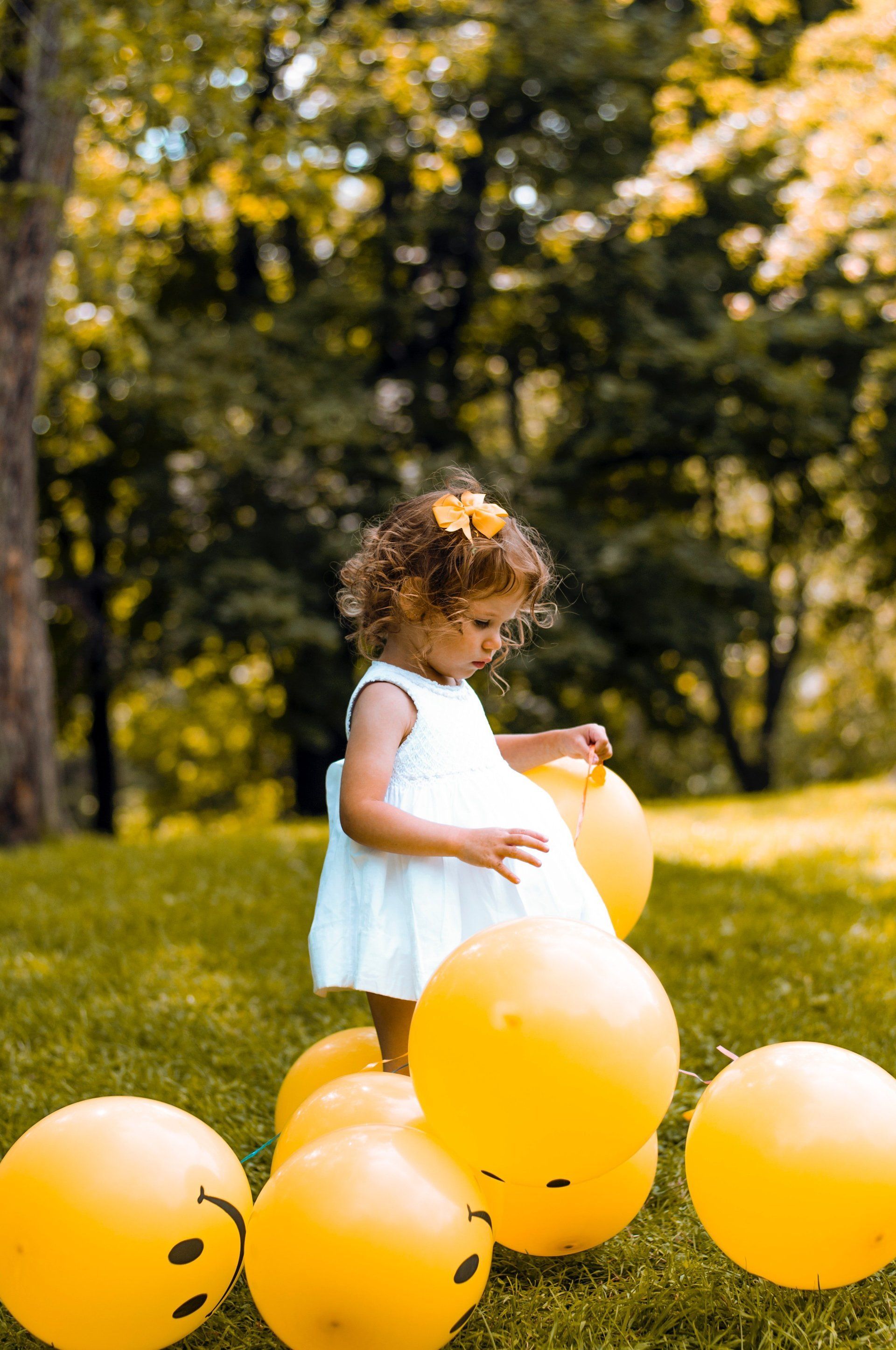 Child in white dress plays with yellow balloons in a grassy park, smiling.