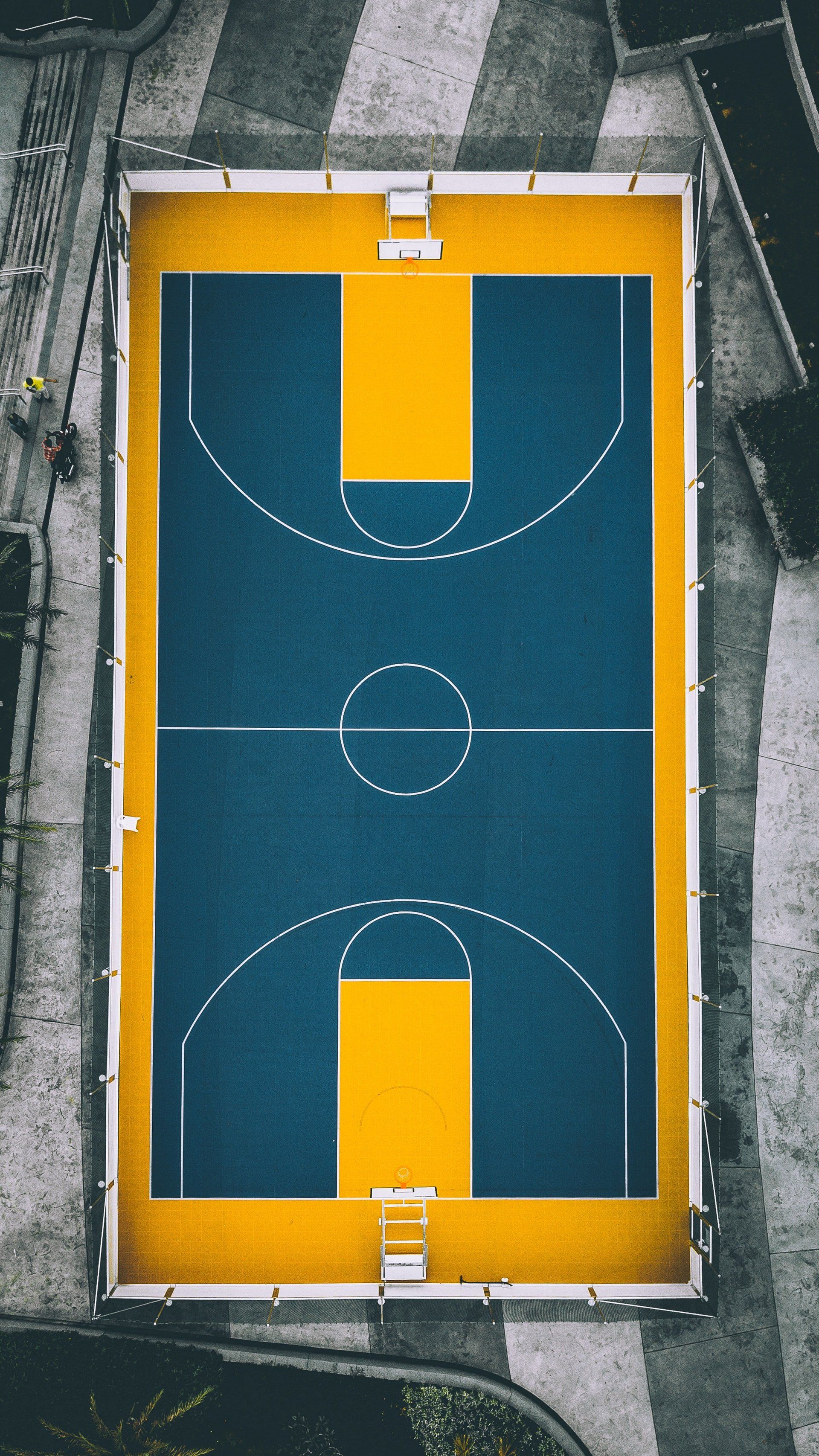 Top-down view of an outdoor blue-and-yellow basketball half court surrounded by pavement.