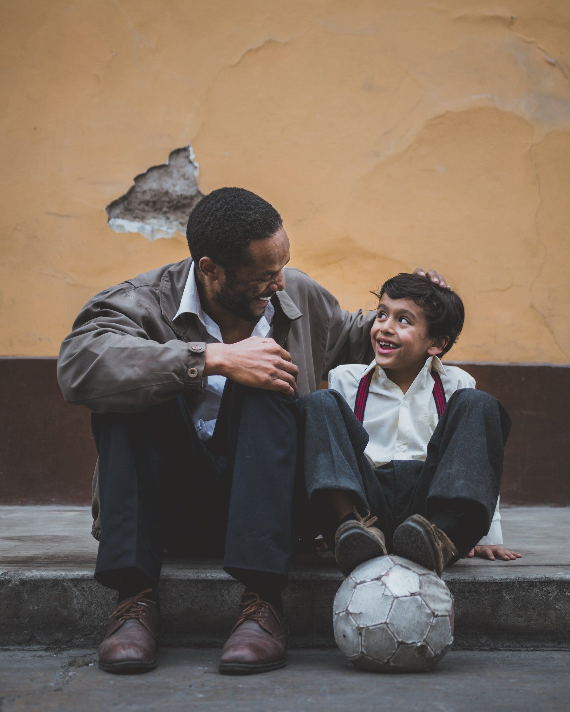 Two people sitting on steps, smiling at each other with a worn soccer ball between them.