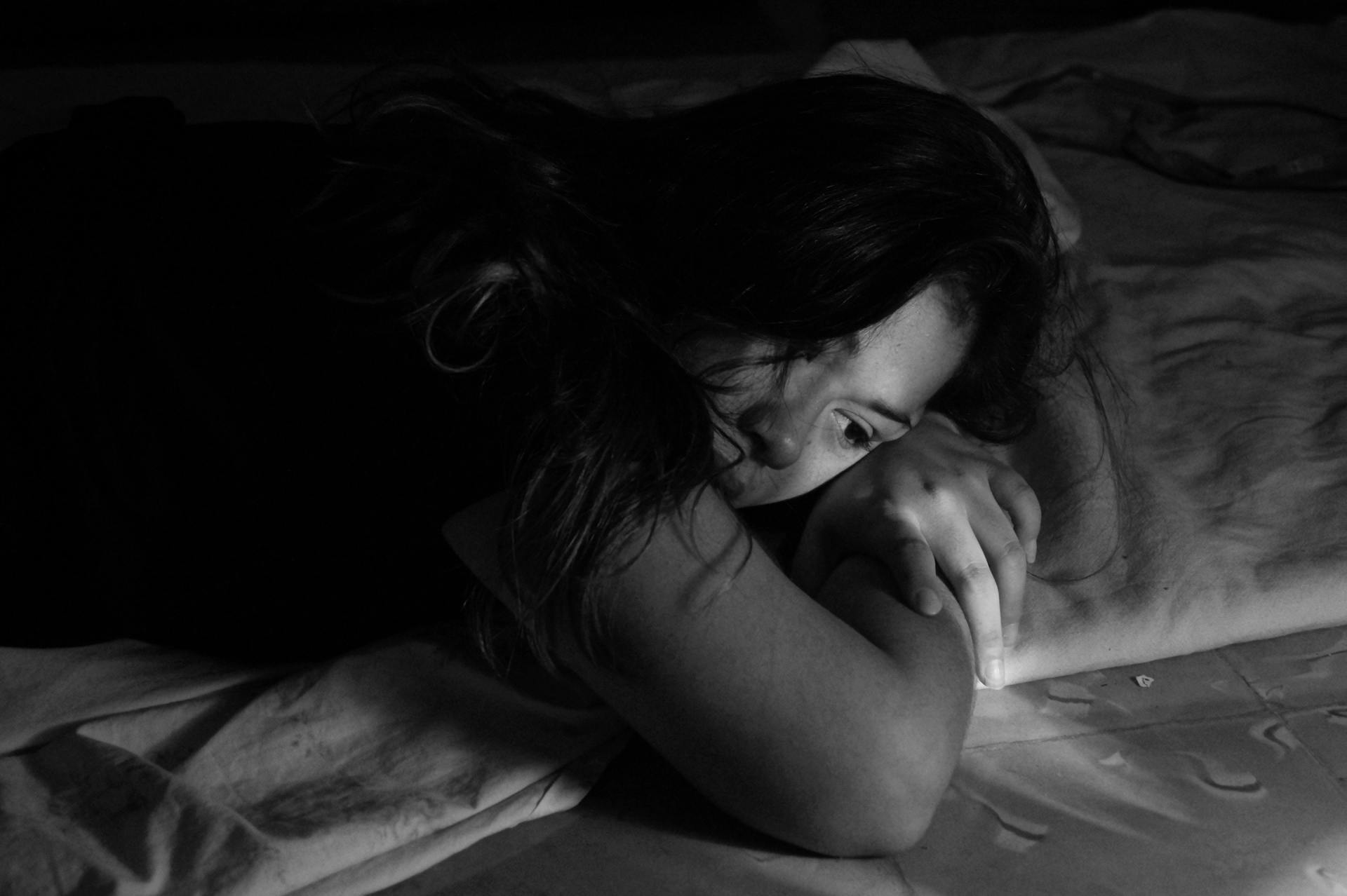 Woman on bed, face down, appears distressed, clutching the sheets, in a dark room.