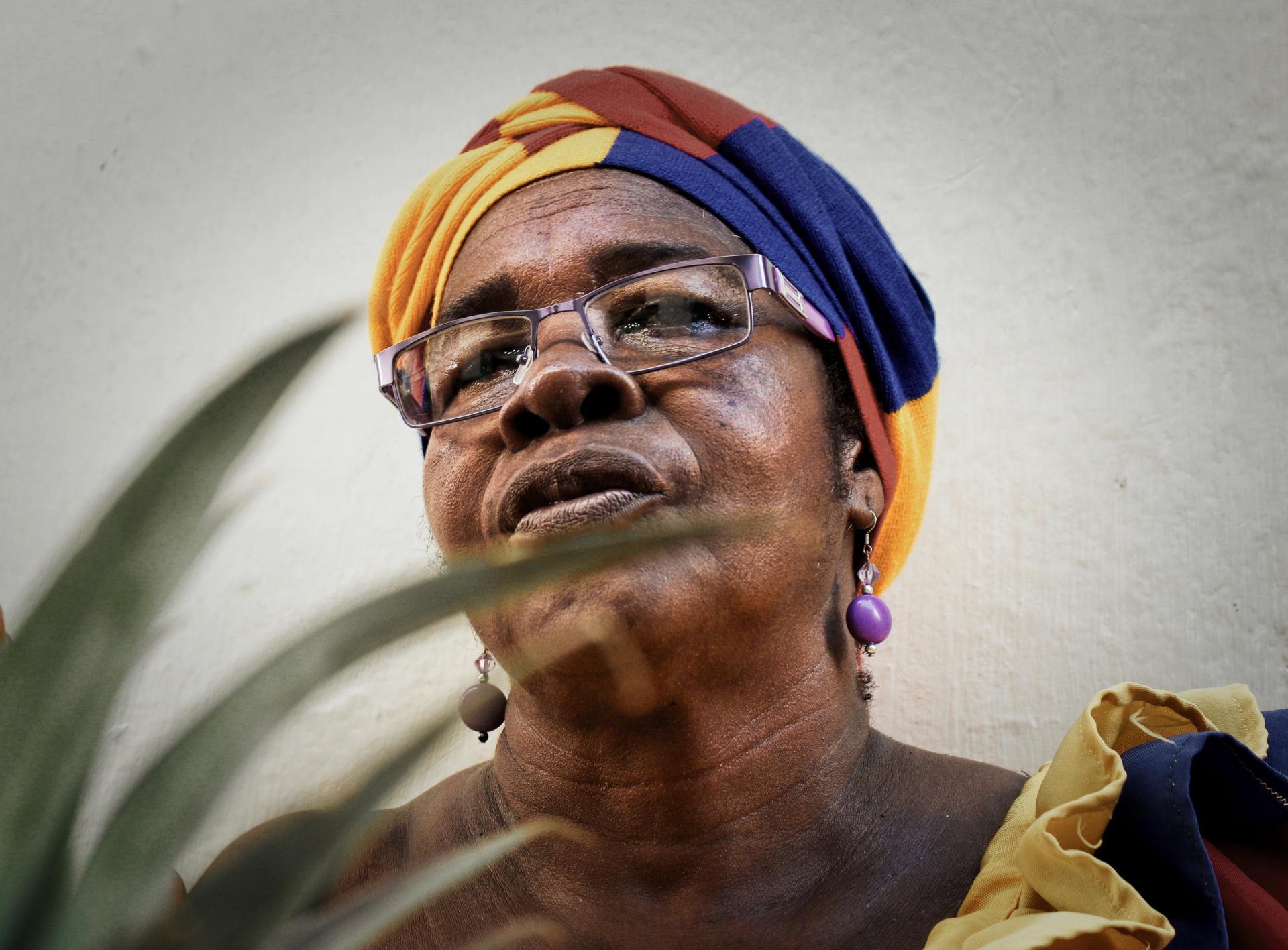 Woman wearing colorful headwrap and glasses, looking up, with a plant in the foreground.