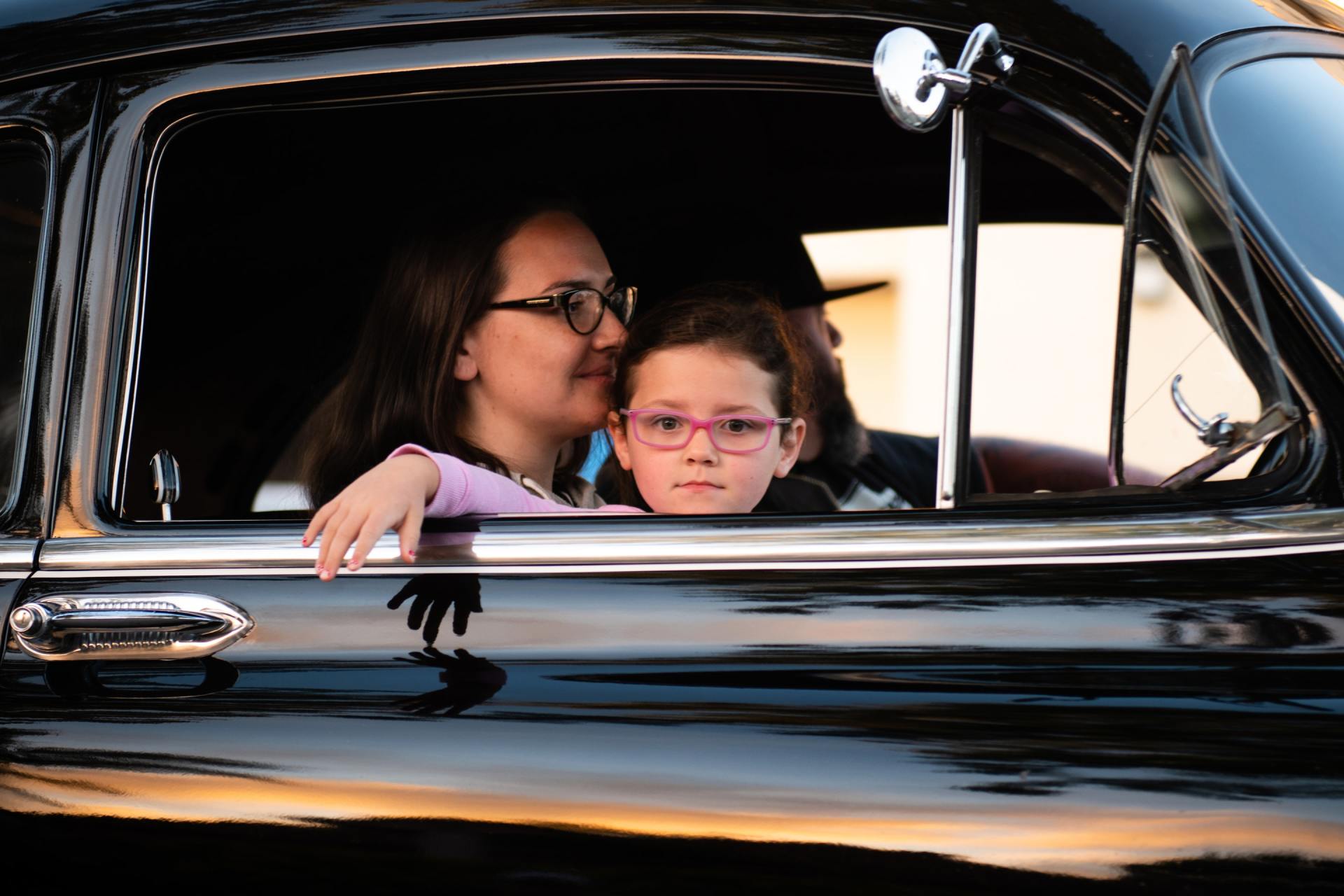 Woman and child look out from a classic black car. Child in pink glasses rests head on woman's shoulder.