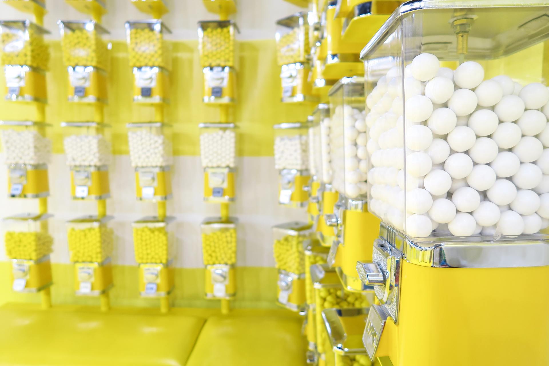 A bunch of gumball machines are lined up against a yellow wall.
