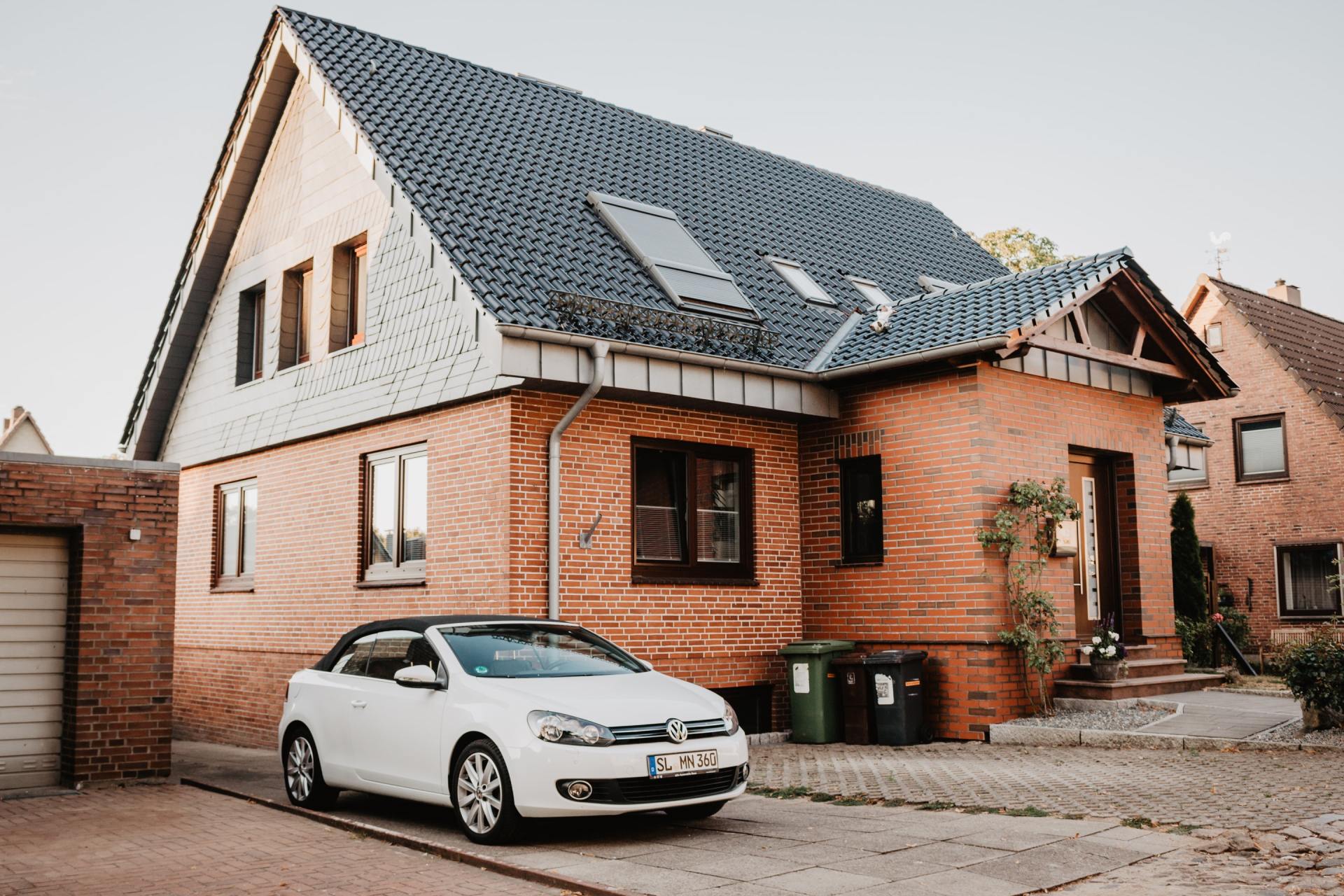 White convertible car parked in front of a brick house with a dark gray roof.