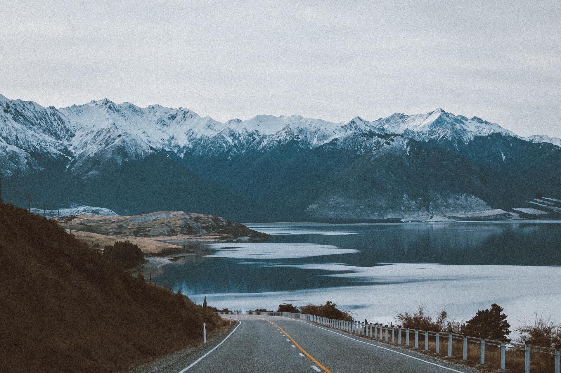 Road leading towards snow-capped mountains and a calm lake under a cloudy sky.