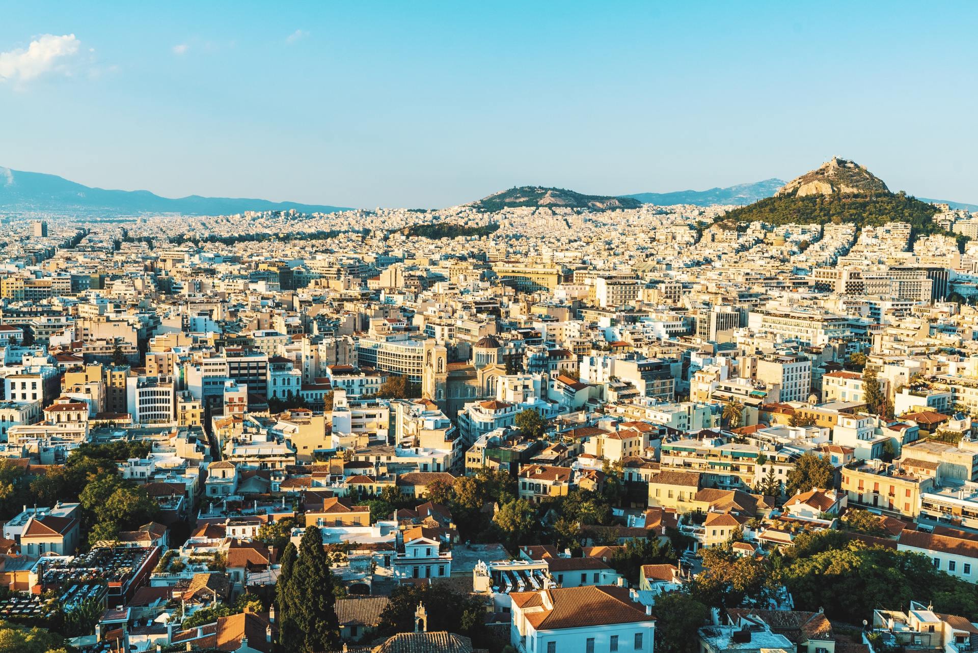 Αεροφωτογραφία της Αθήνας, Ελλάδα. Κτίρια απλώνονται στο τοπίο κάτω από έναν γαλάζιο ουρανό, με μια εξέχουσα κορυφή λόφου στο βάθος.