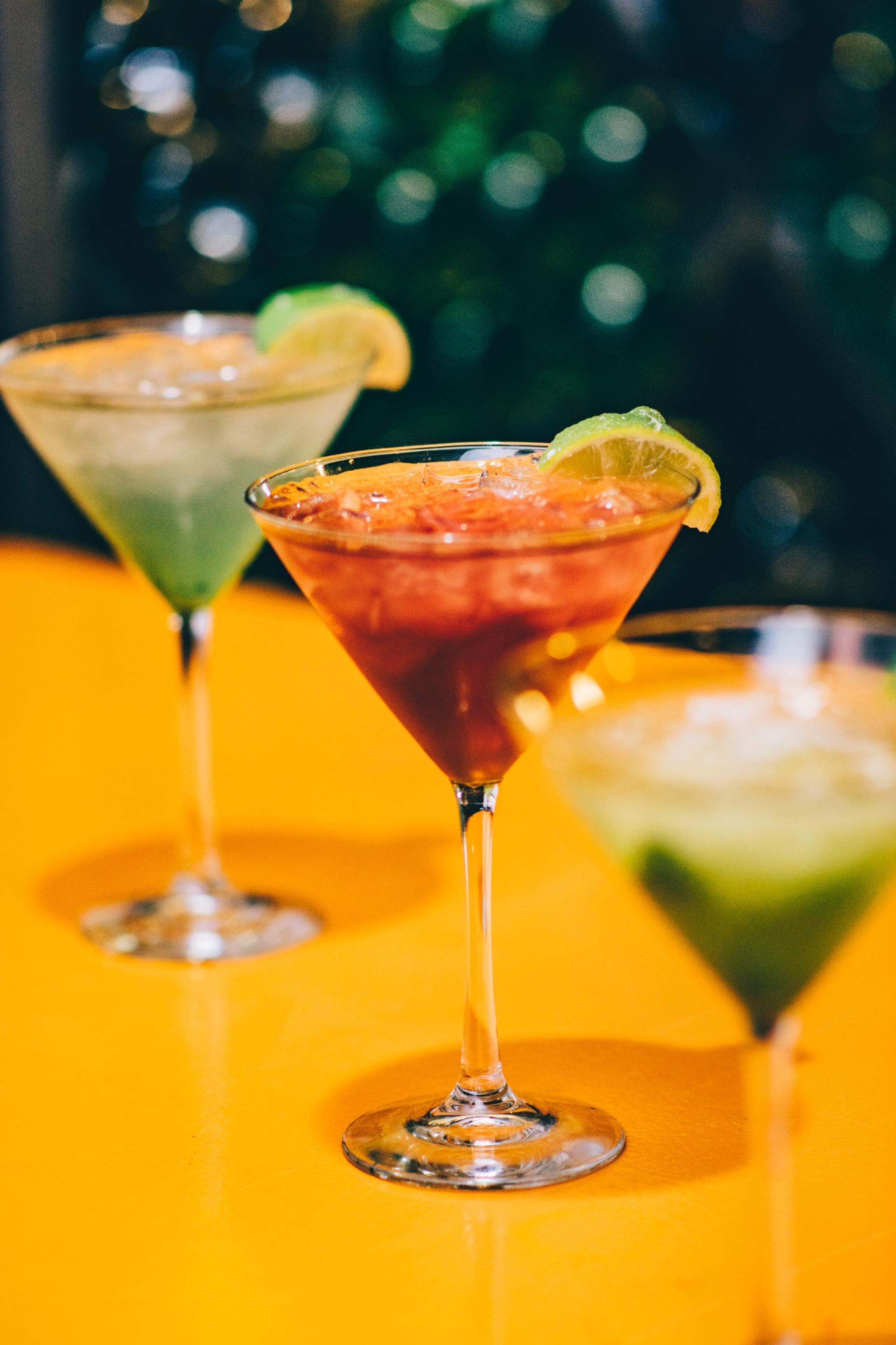 Three colorful cocktails in martini glasses, garnished with lime, on a yellow surface, with a green background.